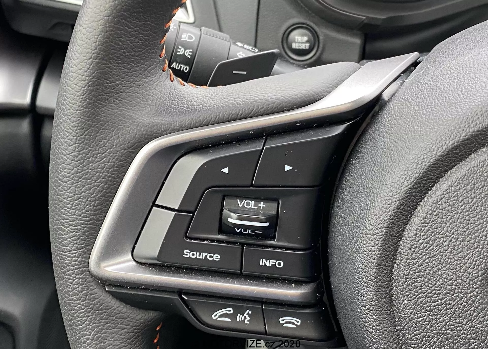 Vista ravvicinata dei comandi del volante della Subaru XV 2018 dalla prospettiva del sedile del conducente. L'immagine mette in risalto i pulsanti multifunzione sullo sparo sinistro, inclusa la regolazione del volume, il pulsante INFO per le impostazioni del display e un'icona che sembra un menu. Gli interruttori del controllo della velocità sono leggermente fuori fuoco in alto. I controlli sono realizzati in plastica e gomma, con un volante rivestito in pelle che presenta un design ergonomico. I metadati nell'angolo in basso a destra recitano 'IZE-EV2020'.