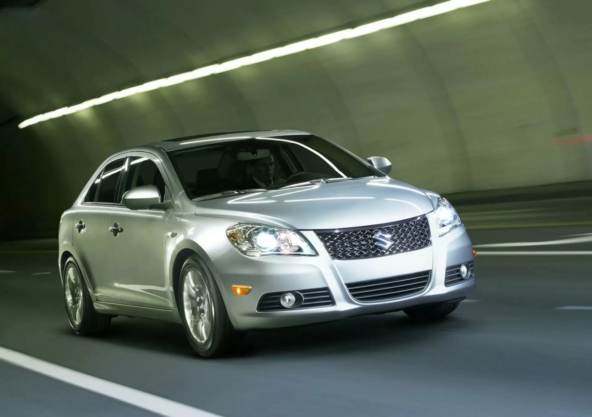 Berlina argento Suzuki Kizashi 2010 in transito in un tunnel, con griglia anteriore, fari LED, luci antinebbia, cerchi in lega e vernice riflettente, vista da un angolo basso frontale di tre quarti che sottolinea il design dinamico e la tecnologia del veicolo.