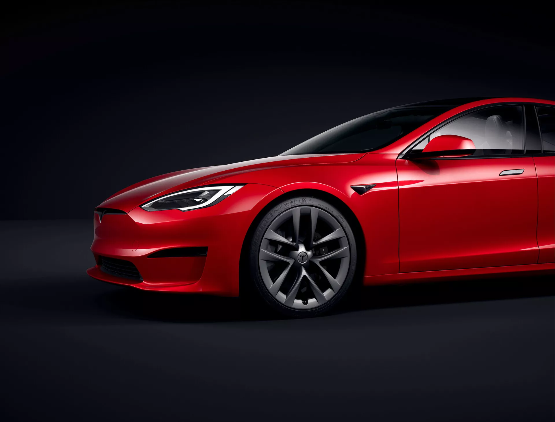 Tesla Model S 2016 (restyling di prima generazione) berlina rossa in studio con illuminazione controllata e sfondo nero, vista angolare bassa che enfatizza il profilo aerodinamico e l'altezza da terra, vista frontale tre quarti mostrando il lato del conducente, fari, griglia, parte del cofano, design dei cerchi in lega premium con motivo a più raggi, parabrezza con vetro oscurato, linea del tetto inclinata, specchietto laterale, maniglie delle porte e linea dei finestrini che si integrano armoniosamente nel design elegante, evidenziando la finitura della vernice, linee di contorno e pannelli scolpiti che suggeriscono lusso e performance, illuminazione focalizzata che mette in risalto i dettagli del design.