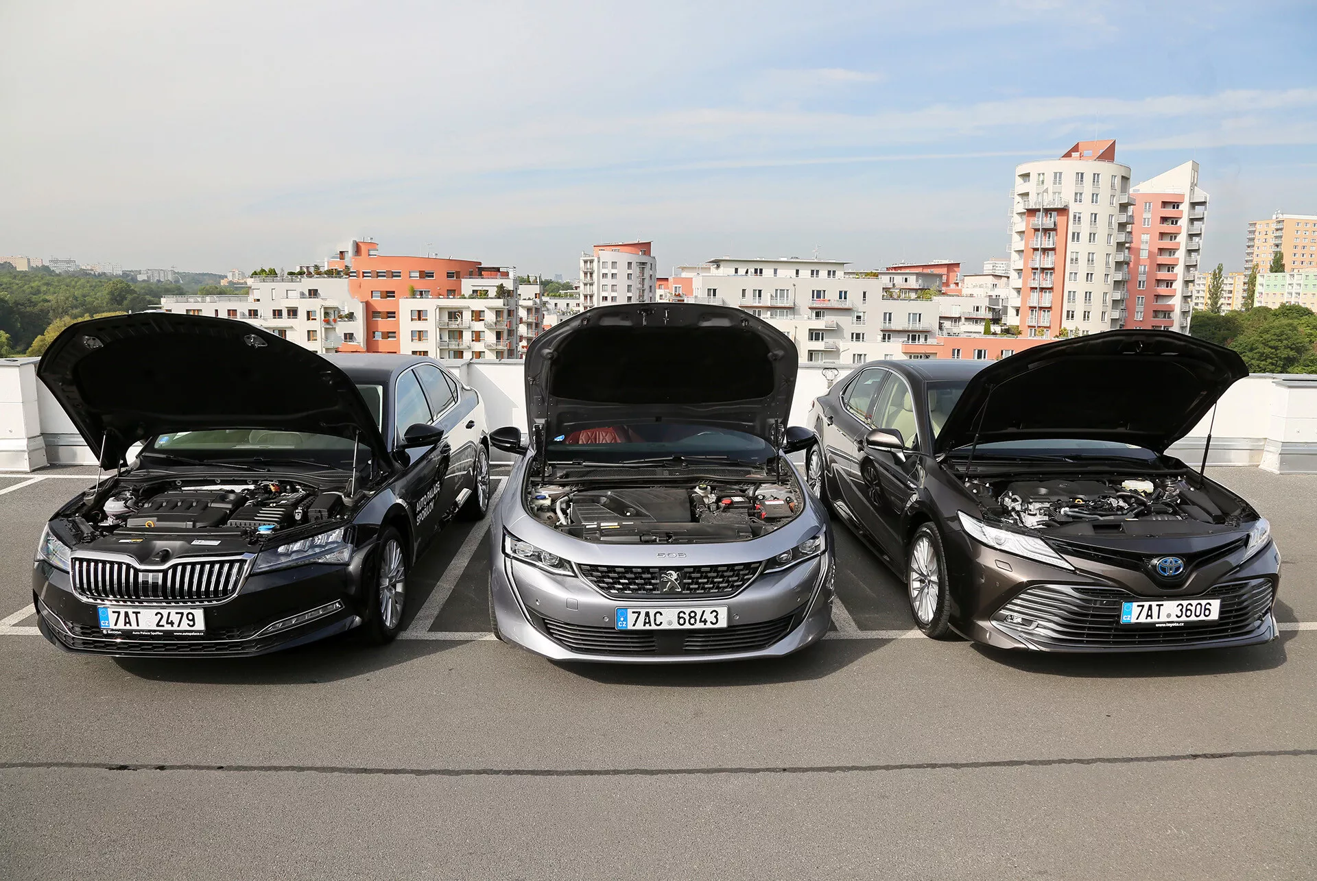 Modelli limousine Toyota Camry 2019 con cofani aperti che mostrano i motori, parcheggiati uno accanto all'altro su una struttura rialzata, con lo skyline della città sullo sfondo, evidenziando il design elegante, moderno dei veicoli e la tecnologia avanzata del motore.