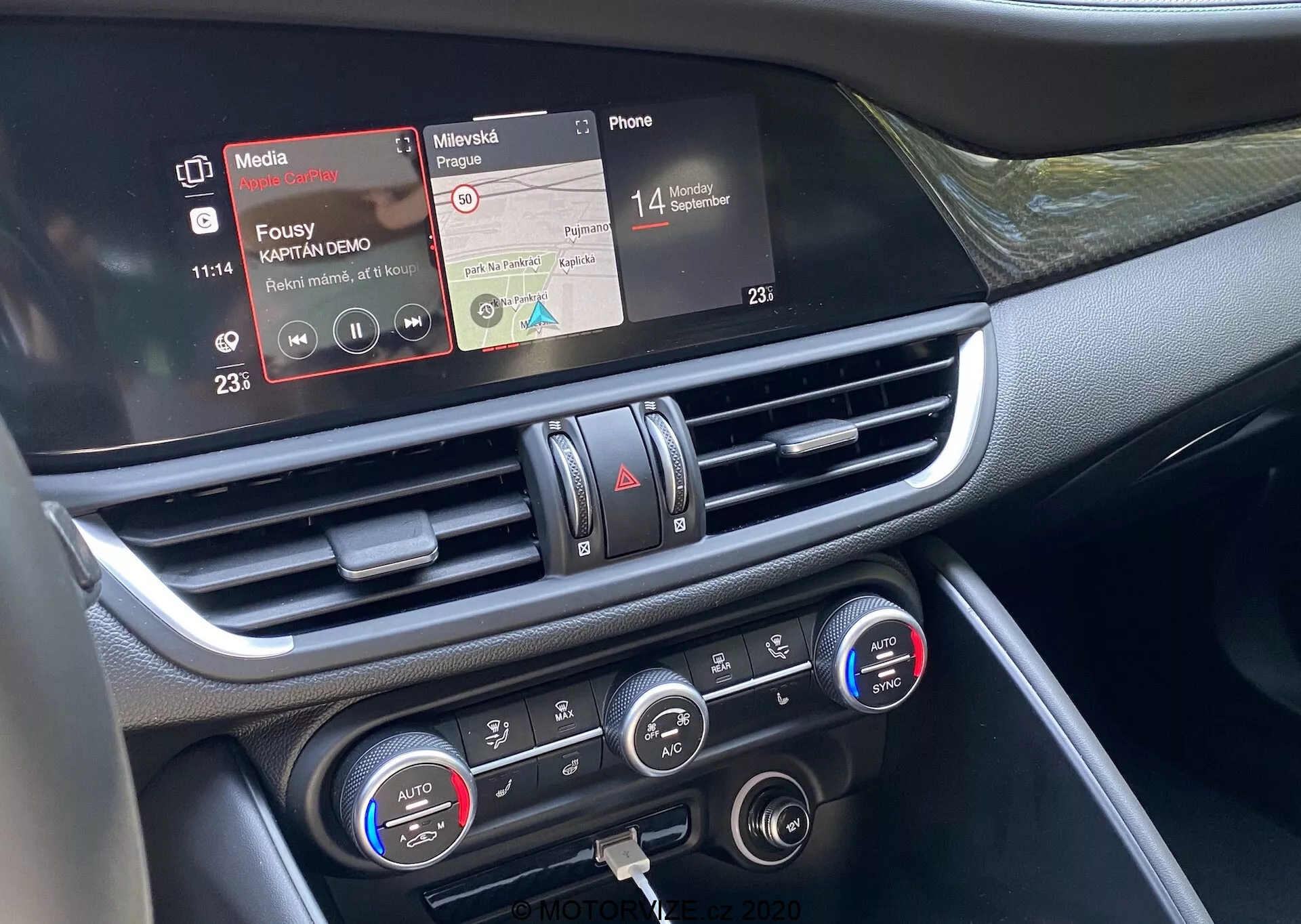 Vista ravvicinata della sezione centrale del cruscotto dell'Alfa Romeo Giulia 2016 dal sedile del passeggero anteriore. L'immagine in primo piano evidenzia il touchscreen dell'infotainment con funzioni di riproduzione multimediale e connettività telefonica, la bocchetta dell'aria centrale con accenti cromati e i comandi del clima con impostazioni della temperatura a doppia zona e display digitali. I materiali del cruscotto sembrano includere plastica e rifiniture metalliche o cromate, mostrando la tecnologia avanzata degli interni del veicolo focalizzata sulla connettività, riproduzione multimediale e controllo climatico.