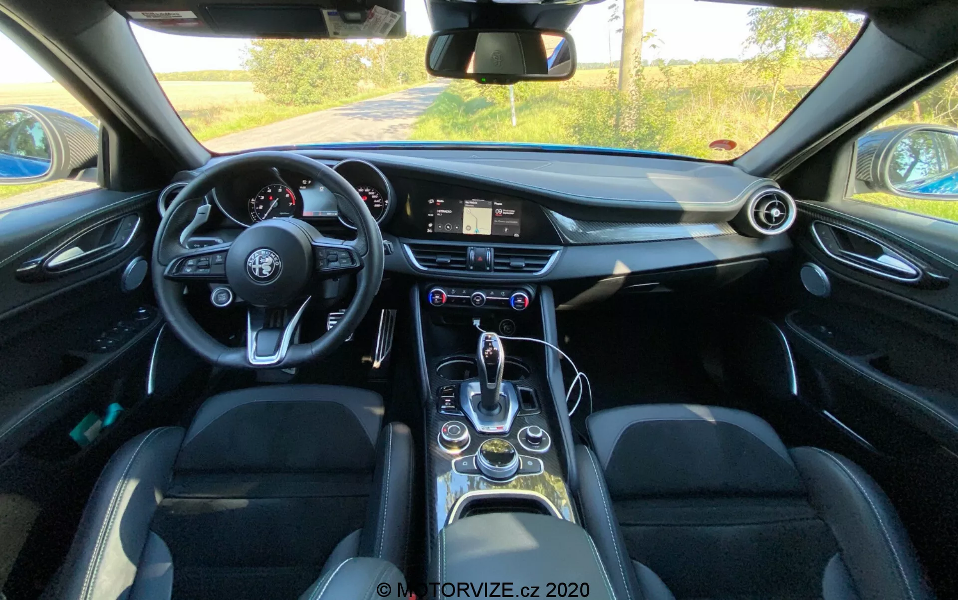 Vista interna di una Alfa Romeo Giulia 2016 dalla prospettiva del guidatore che mostra un volante a fondo piatto con più pulsanti di controllo, un sistema di infotainment digitale sul cruscotto, un orologio analogico, prese d'aria circolari, una leva del cambio elegante, selettore delle modalità di guida, portabicchieri, finestre elettriche, sedili in pelle con supporti laterali e caratteristiche tecnologiche moderne in generale.