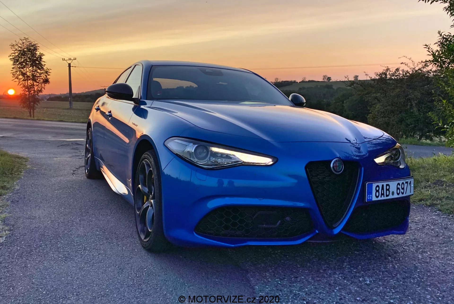 Una berlina sportiva Alfa-Romeo Giulia 2016 blu catturata di sera con un tramonto sullo sfondo, posizionata leggermente angolata verso la fotocamera su una strada di campagna pavimentata, che mostra la sua distintiva griglia anteriore, il design aggressivo dei fari a LED, grandi cerchi in lega multirazze e un sistema frenante ad alte prestazioni. La vernice metallizzata, il paraurti anteriore aerodinamico, le linee scolpite del cofano e i sensori avanzati di assistenza alla guida evidenziano l'estetica sportiva e la tecnologia moderna della vettura. È presente una filigrana 'MOTORVIZE.cz 2020'.