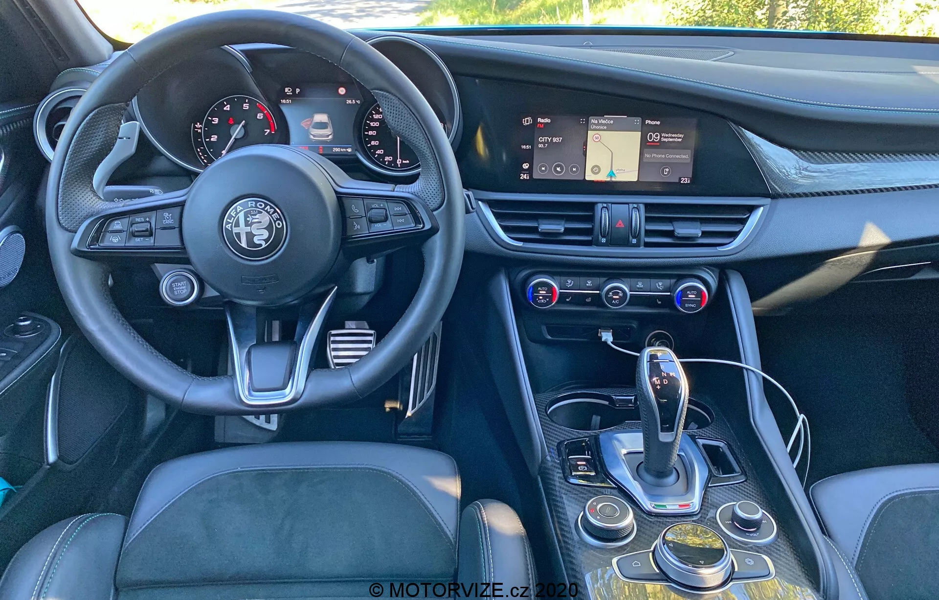 Vista interna di un'Alfa Romeo Giulia 2016 da un angolo leggermente decentrato, da dietro verso il fronte, che mostra il cruscotto, il volante, la console centrale e parte del sedile del conducente. L'immagine mette in risalto il volante a tre razze con l'emblema Alfa Romeo, un quadro strumenti con quadranti analogici e display digitali, un cruscotto con un sistema di infotainment touchscreen, prese d'aria e controlli per il clima e i multimedia. La console centrale include un selettore di cambio automatico, un quadrante rotante, pulsanti per la navigazione dell'infotainment e le impostazioni del veicolo e un possibile selettore di modalità. Il design enfatizza l'ergonomia e l'accessibilità, con materiali come pelle, metallo spazzolato e plastica di alta qualità per una sensazione di lusso.