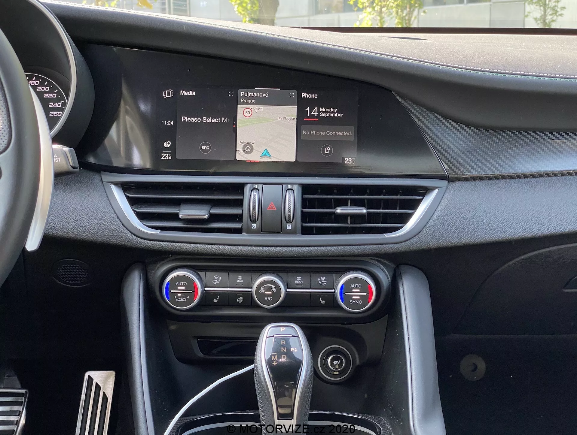 Vista interna della Alfa-Romeo Giulia 2016 con focus sulla plancia centrale con uno schermo infotainment che mostra una mappa di navigazione, prese d'aria orizzontali, pannello di controllo del climatizzatore a doppia zona con manopole e pulsanti, e la leva del cambio parzialmente visibile con rivestimento in pelle e accenti cromati, rappresentando l'ergonomia e la tecnologia degli interni delle auto moderne.