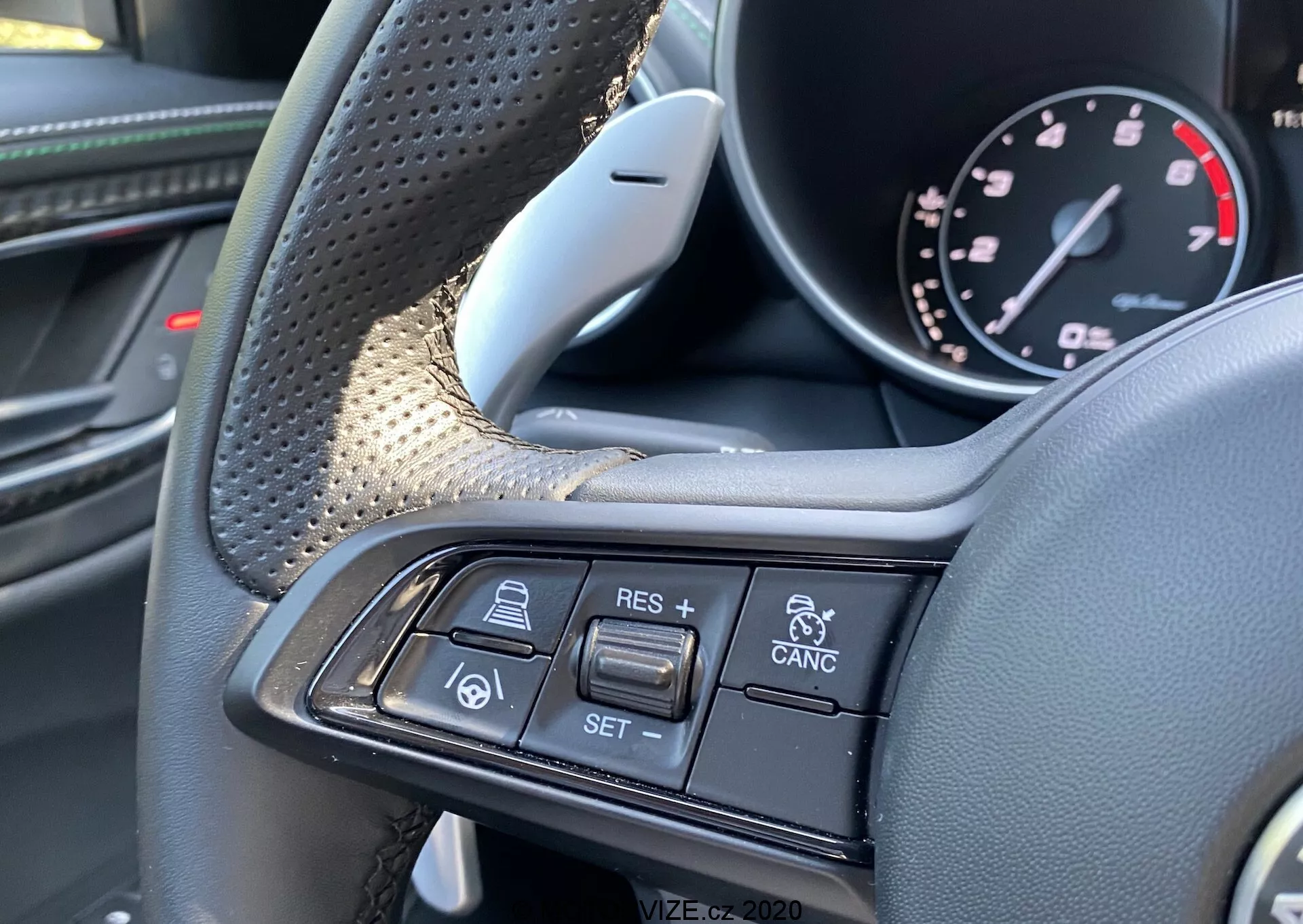 Vista ravvicinata degli interni dell'Alfa-Romeo Giulia 2016 che mostra il volante e i suoi comandi multifunzione dalla prospettiva del conducente. Le aree di presa testurizzate e i materiali lussuosi come la pelle e forse la fibra di carbonio sono visibili sul lato sinistro. Il raggio destro presenta i pulsanti del controllo della velocità di crociera e della tecnologia di controllo adattivo, tra cui + RES, - SET, CANCEL, un'icona di auto che segue un'altra auto e avvisi di collisione dei veicoli. L'immagine si concentra esclusivamente su questi controlli, enfatizzando il design premium e le funzioni avanzate di assistenza alla guida.
