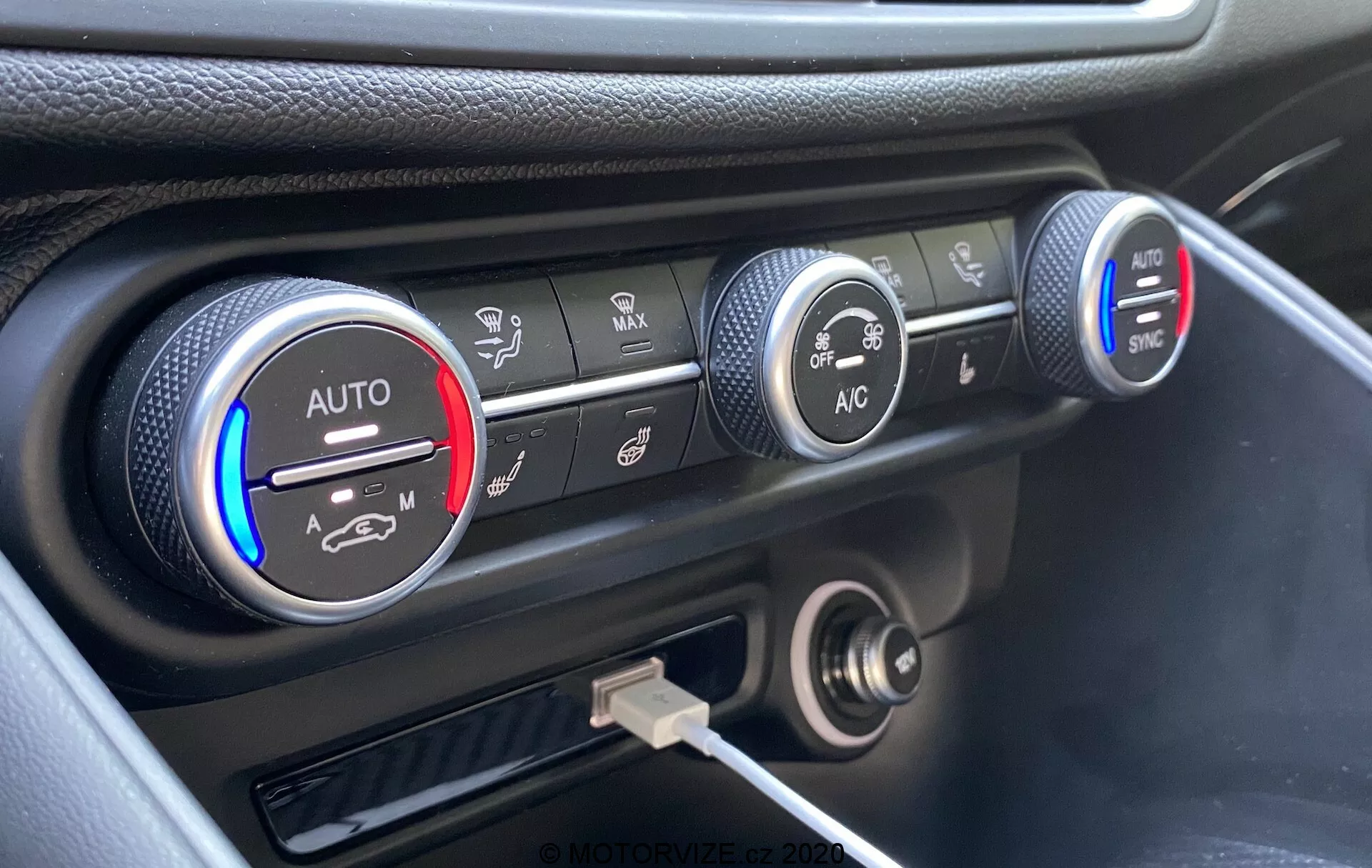Primo piano del sistema di controllo del clima sul cruscotto della Alfa Romeo Giulia 2016, con tre manopole circolari. La manopola sinistra indica le impostazioni della temperatura con bande blu e rosse e una impostazione 'AUTO', parzialmente illuminata in rosso. La manopola centrale mostra le impostazioni della velocità della ventola, evidenziate a una velocità inferiore. La manopola destra mostra varie modalità di distribuzione dell'aria con pulsanti 'AC' e 'SYNC'. Sotto le manopole c'è una presa di corrente a 12 volt e una porta USB. Il cruscotto ha una finitura nera con un accento argentato intorno ai controlli del clima, catturato da un angolo elevato.