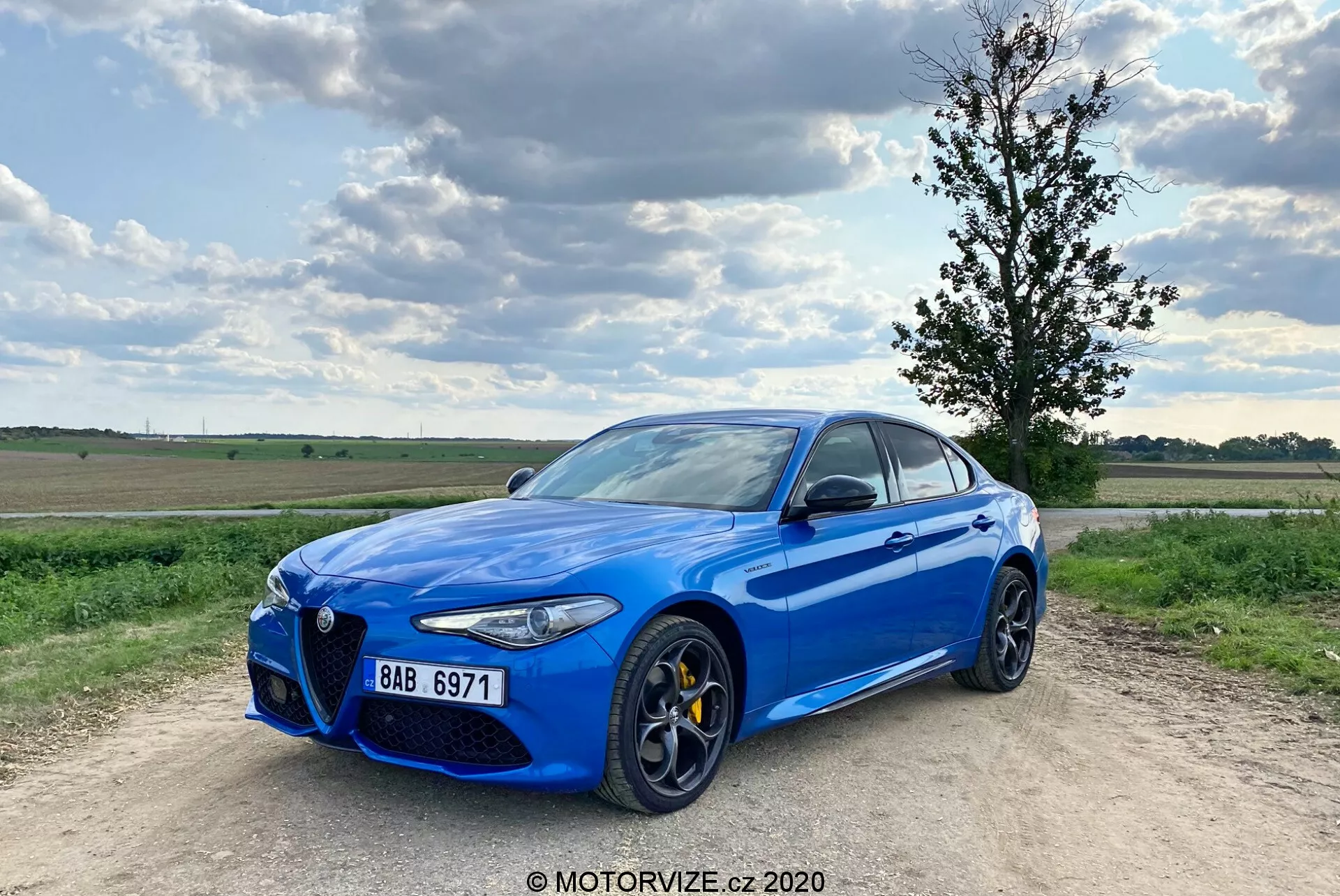 Immagine di una Alfa Romeo Giulia 2016 berlina sportiva blu brillante parcheggiata su un sentiero sterrato, con un cielo parzialmente nuvoloso sullo sfondo. L'auto è ripresa da un angolo frontale a tre quarti, mostrando la sua distintiva griglia frontale 'Trilobo', pinze dei freni gialle, grandi cerchioni in lega a più razze, eleganti fari a LED e cornici dei finestrini cromate. Il veicolo è leggermente fuori centro verso sinistra con la ruota anteriore sinistra puntata verso l'esterno, conferendogli una posizione dinamica.