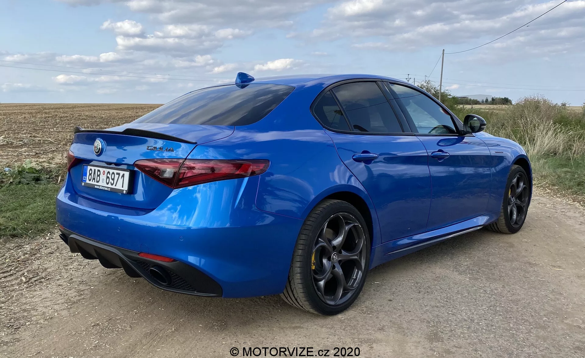Vista tre quarti posteriore di una berlina sportiva blu brillante su una strada non asfaltata con campo aperto e cielo parzialmente nuvoloso sullo sfondo. Fotografia a bassa angolazione che mette in risalto la postura dell'auto, paraurti posteriore scolpito con doppi scarichi integrati, gruppi ottici posteriori sottili e orientati orizzontalmente, spoiler sul cofano del bagagliaio e cerchi in lega neri a più razze con pneumatici ribassati. Il profilo laterale mostra una silhouette elegante con una linea del tetto inclinata e finestrini posteriori leggermente oscurati. La parte posteriore dell'auto presenta un badge e un emblema del marchio che indicano il modello o il livello di allestimento. L'esterno dell'auto sembra essere in eccellenti condizioni e la luce del giorno offre una chiara visibilità del design e della verniciatura dell'auto.