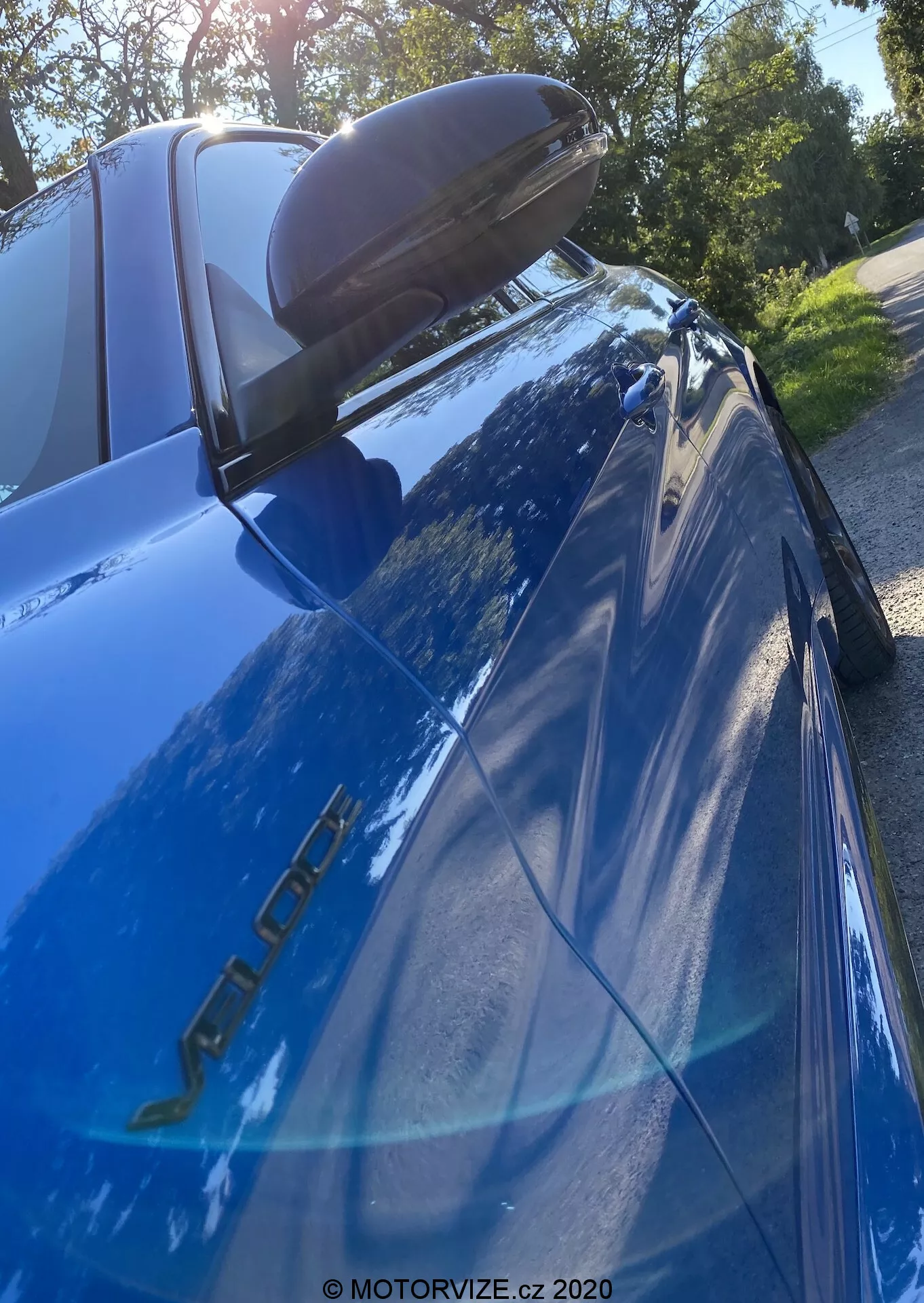 Immagine ravvicinata di una Alfa Romeo Giulia blu (modello 2016) fotografata da un angolo basso e verso l'alto con messa a fuoco sulla sezione posteriore. Sono visibili le targhette del modello sul lato del conducente, la vernice blu riflettente, la luce posteriore a LED sinistra, lo spoiler posteriore nero, il finestrino posteriore inclinato con la terza luce di stop, il riflesso di alberi e cielo luminoso e lo specchietto laterale con indicatore di direzione integrato.