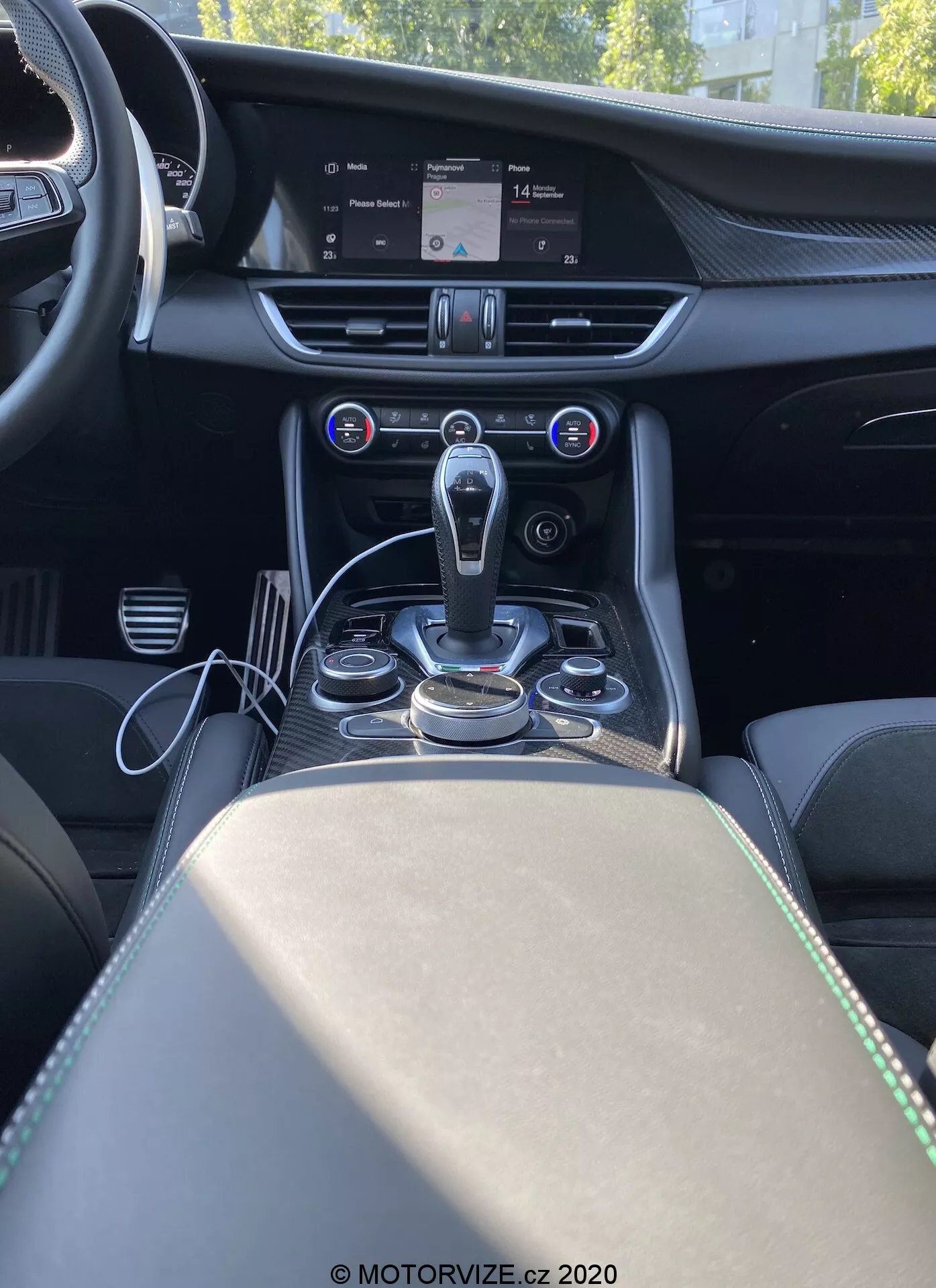 Scatto interno dell'Alfa Romeo Giulia 2016 visto da dietro la console centrale, mostrando la plancia centrale, il tunnel della trasmissione e parte del sedile del passeggero. L'immagine offre una prospettiva frontale dalla visuale di un passeggero posteriore, evidenziando una moderna leva del cambio automatico circondata da manopole di controllo e pulsanti metallici per il sistema di infotainment e le modalità di guida. Al di sopra dell'area del cambio si trova il sistema di climatizzazione automatica bi-zona con un display digitale che mostra le impostazioni della temperatura e pulsanti per controllare la distribuzione dell'aria e la velocità della ventola. Un ampio schermo di infotainment in cima alla plancia centrale mostra una mappa del sistema di navigazione. Il cruscotto presenta dettagli cuciti e accenti metallizzati che suggeriscono materiali premium. Il bracciolo e il vano portaoggetti con due portabicchieri sono visibili dietro il cambio, arricchendo il design lussuoso e orientato alla tecnologia dell'abitacolo.