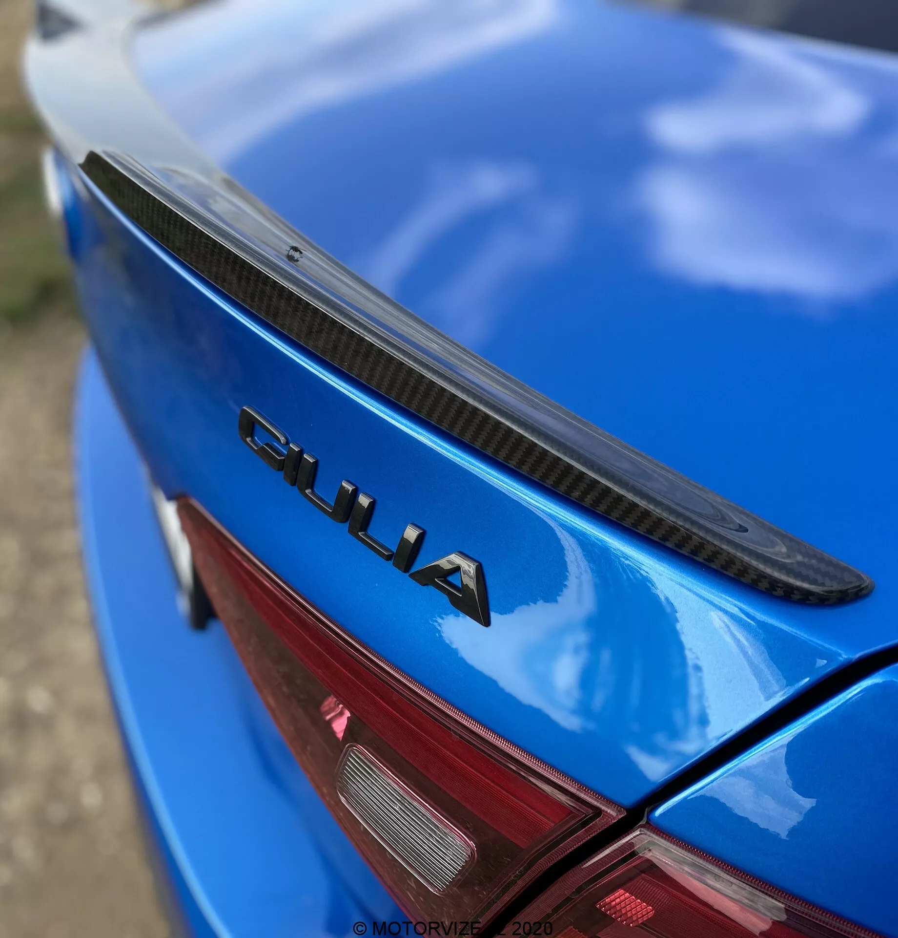 Vista ravvicinata e angolata della sezione posteriore di una Alfa Romeo Giulia 2016 blu. La fotografia mostra il portellone del bagagliaio con uno spoiler nero, vernice blu lucida e un prominente badge cromato 'GIULIA'. È visibile anche una parte della luce di coda LED rossa. Il badge 'GIULIA' è nitidamente a fuoco, mentre lo spoiler, la superficie dell'auto e la luce di coda si sfumano gradualmente, enfatizzando gli elementi di branding e design dell'auto.