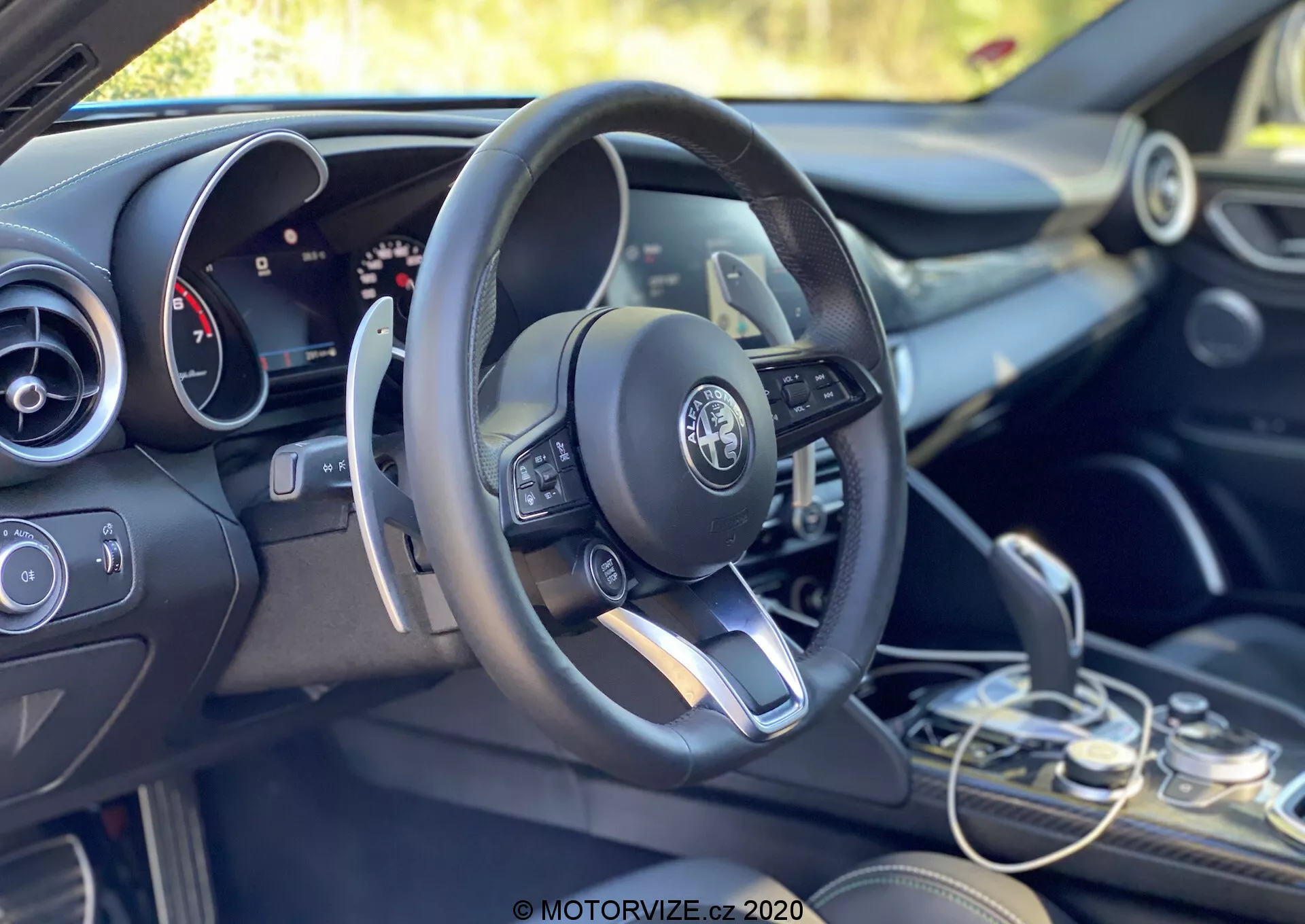Vista interna di una Alfa Romeo Giulia 2016 concentrata sulla zona del conducente e sul cruscotto da una prospettiva leggermente inclinata verso il retro dalla parte sinistra. La configurazione a guida a sinistra presenta un volante multifunzione moderno con comandi integrati, inclusi pulsanti per regolare il volume, accedere ai menu e probabilmente il cruise control. Il logo Alfa Romeo è chiaramente visibile al centro del volante. Dietro il volante, parzialmente visibile, un pannello strumenti misto di display analogici e digitali suggerisce un sistema di informazioni per il conducente configurabile. Alla destra del volante, è appena visibile la parte superiore dello schermo del sistema di infotainment, affiancato sotto dai comandi del climatizzatore e dagli interruttori funzionali. Il selettore del cambio si trova nell'angolo in basso a destra, mostrando un design elegante di trasmissione automatica. Le rifiniture combinate di tonalità scure e accenti metallici riflettono il design interno premium del veicolo. L'abitacolo è inondato di luce naturale, con uno sfondo verde che suggerisce un'ambientazione esterna o vegetata.