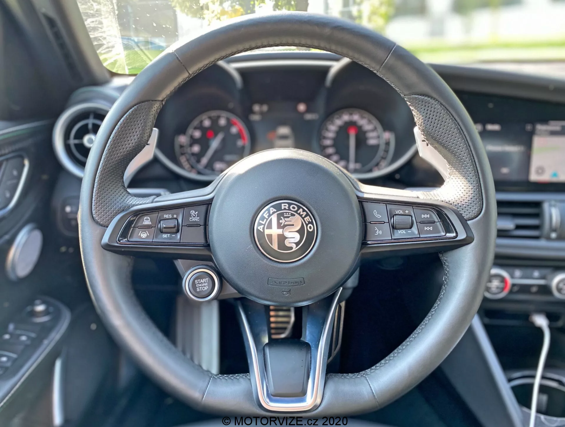 Prospettiva del conducente dell'interno dell'Alfa-Romeo Giulia 2016 con focus sul volante e sul cruscotto. Il volante posizionato centralmente ha un design multifunzionale con pulsanti per media, controllo vocale, connettività telefonica, informazioni del veicolo e impostazioni del cruise control. Il quadro strumenti mostra quadranti analogici, probabilmente un tachimetro e un contagiri, con un display digitale tra di loro. Il volante combina materiali come pelle, cuciture e accenti metallici, emanando lusso. Gli elementi sullo sfondo includono i comandi del climatizzatore e uno schermo centrale sopra di essi.