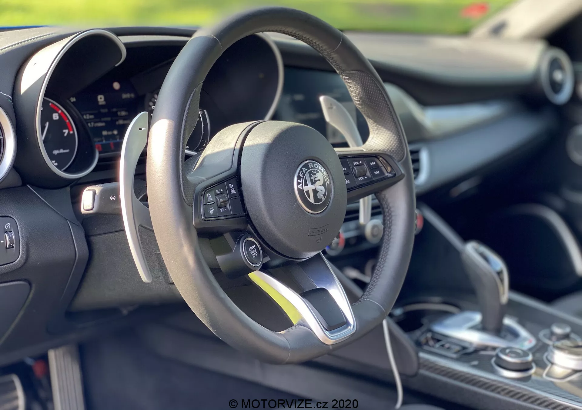 Scatto degli interni di un Alfa-Romeo Giulia 2016 che si concentra sull'area del conducente e sul volante. Il volante piatto in basso rivestito in pelle presenta comandi multifunzione sul lato sinistro. Dietro il volante è visibile un cruscotto digitale. La console centrale mostra una leva del cambio con un rivestimento in pelle, suggerendo possibili capacità di cambio manuale. L'immagine enfatizza il design sportivo dell'auto, la tecnologia avanzata e l'artigianato dettagliato con una bassa profondità di campo che mette in evidenza gli elementi in primo piano.