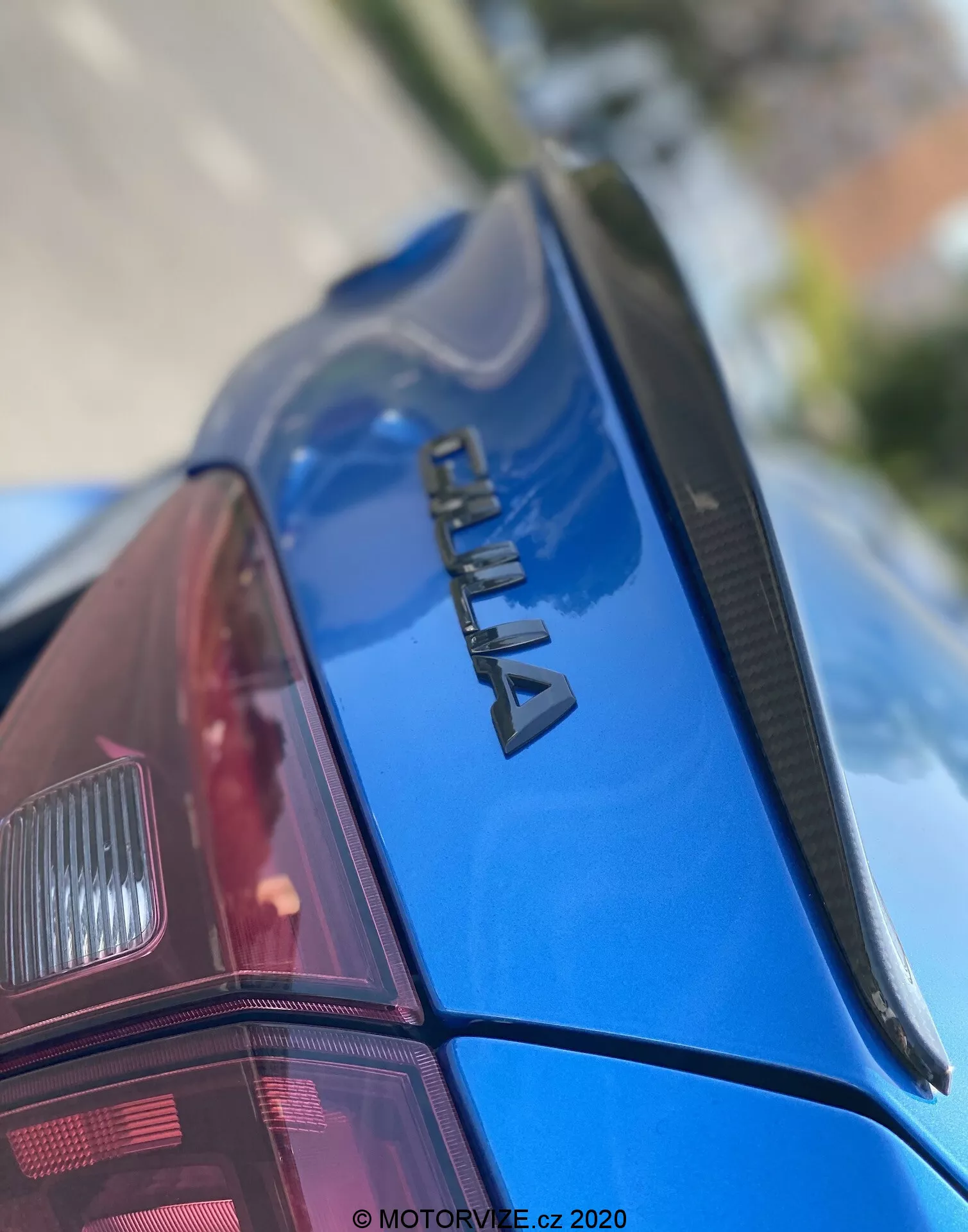 Primo piano della sezione posteriore di un'auto blu ŠKODA, vista da una prospettiva verso l'alto. L'attenzione è sul logo cromato 'ŠKODA' sul cofano del bagagliaio, uno spoiler sottile e la vernice lucida e ben mantenuta. Il parabrezza posteriore ha linee di sbrinamento e si intravede parzialmente il gruppo ottico posteriore di design moderno. La scarsa profondità di campo enfatizza il marchio, con la parte anteriore e lo sfondo sfocati. Con filigrana 'MOTORVIZE.cz 2020', indicando la data di pubblicazione o acquisizione.
