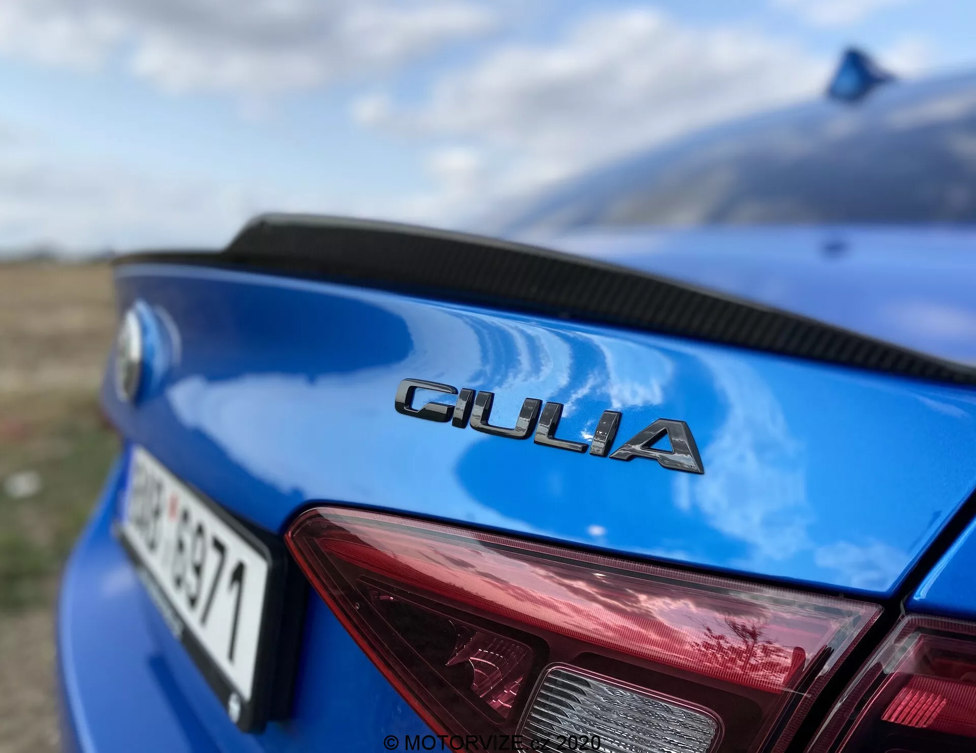 Primo piano della parte posteriore di un Alfa Romeo Giulia 2016 blu, ripresa da un'angolazione bassa concentrandosi sul badge cromato 'GIULIA' mostrato in modo prominente sul coperchio del bagagliaio. Le lettere nitide contrastano con la vernice blu, evidenziando la tipografia del marchio. In alto a destra è visibile una porzione segmentata della luce posteriore destra, che mostra un design rosso distintivo con accenti bianchi per le luci di retromarcia e gli indicatori di direzione. La targa è parzialmente visibile e sfocata sotto la designazione del modello. La curvatura del bagagliaio suggerisce un design aerodinamico, con un paesaggio naturale sfocato sullo sfondo e un effetto bokeh da un’apertura alta, enfatizzando i dettagli del veicolo.