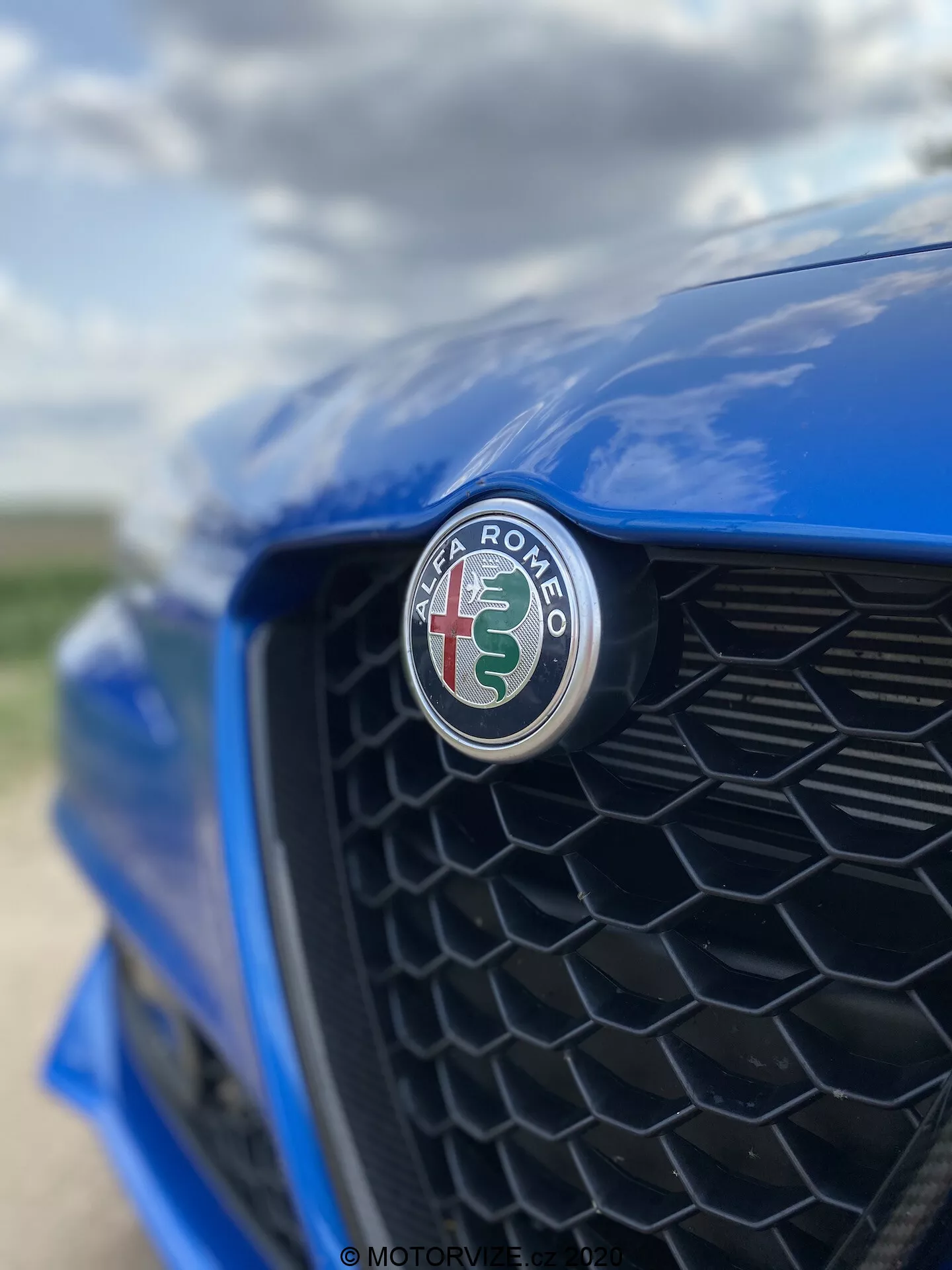 Primo piano della sezione frontale di una Alfa Romeo Giulia 2016 blu, con particolare attenzione al distintivo del produttore con una croce rossa e un serpente verde su sfondo bianco. Scattata da un'angolazione decentrata verso il lato del guidatore, la griglia a nido d'ape nera è leggermente sfocata, evidenziando la messa a fuoco nitida del distintivo e la finitura lucida.