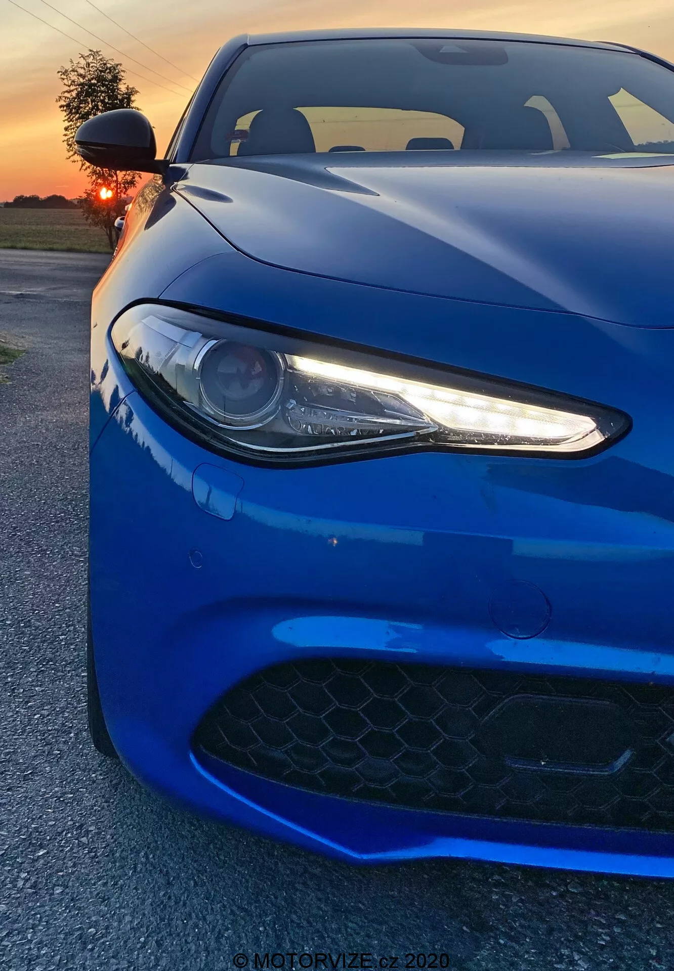 L'immagine ritrae una Alfa Romeo Giulia 2016 sportiva catturata da un angolo frontale basso. Il vibrante veicolo blu intenso riempie l'inquadratura, enfatizzando il faro sinistro con luci diurne a LED, una parte del cofano e la griglia di presa d'aria nel paraurti anteriore. Il faro di tipo a proiettore, con i suoi eleganti accenti LED bianchi, conferisce un aspetto aggressivo e moderno. La griglia nera a trama esagonale suggerisce capacità ad alte prestazioni. La ripresa cattura le linee e i contorni aerodinamici della vettura, con riflessi al tramonto che esaltano la sua finitura lucida. Lo sfondo mostra un cielo arancione illuminato parzialmente oscurato dal sole. L'angolo ravvicinato del fotografo mette in risalto i dettagli del design, evocando una sensazione di potenza ed eleganza.