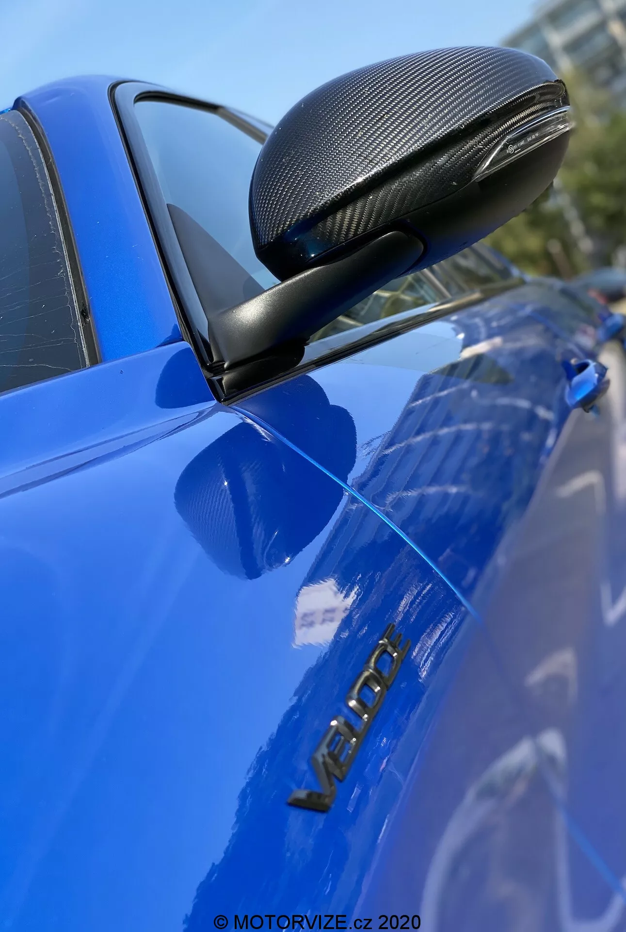 Immagine ravvicinata di una Alfa Romeo Giulia 2016 blu, che enfatizza lo specchietto retrovisore laterale con finitura in fibra di carbonio in una carenatura aerodinamica, montato su un supporto nero. La foto si concentra nitidamente sullo specchietto, sulla porta del conducente e sulla targhetta metallica 'VERON', evidenziando questi dettagli con una ridotta profondità di campo e una superficie lucida e ben curata.