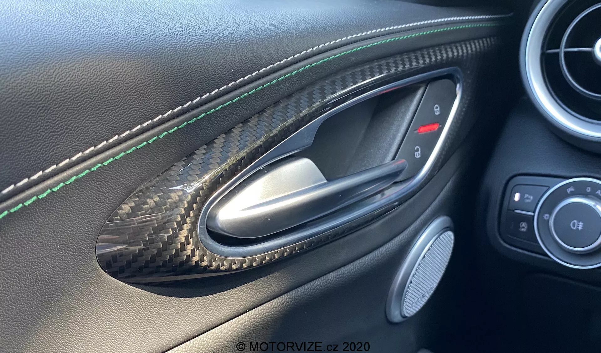 Immagine dell'interno della portiera lato guidatore della Alfa Romeo Giulia 2016 vista dalla prospettiva di una persona seduta all'interno, evidenziando una maniglia della portiera in metallo inserita in una rientranza con motivo in fibra di carbonio, cuciture verdi sul pannello della portiera in pelle, pulsanti di controllo per specchietti laterali e finestrini, griglia dell'altoparlante e finiture in pelle di alta qualità e fibra di carbonio indicativi di un veicolo di lusso o sportivo.