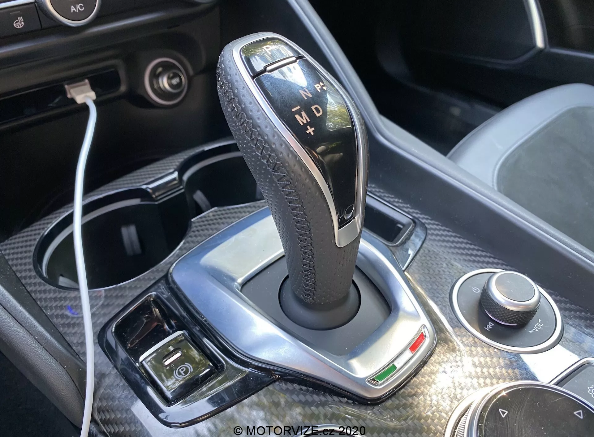 Primo piano della leva del cambio nella trasmissione automatica dell'Alfa-Romeo Giulia 2016, con una cornice argentata lucida, impugnatura nera con texture in pelle e indicatori illuminati per 'P', 'R', 'N', 'D' e 'M'. La luce diurna brillante mette in risalto il design, mostrando un quadrante rotante e connettività USB nelle vicinanze. Finitura cromata, luce indicatrice verde e pulsanti aggiuntivi per freno di stazionamento elettronico e controllo del clima sono visibili, offrendo uno sguardo sul lusso e la tecnologia avanzata degli interni dell'auto.