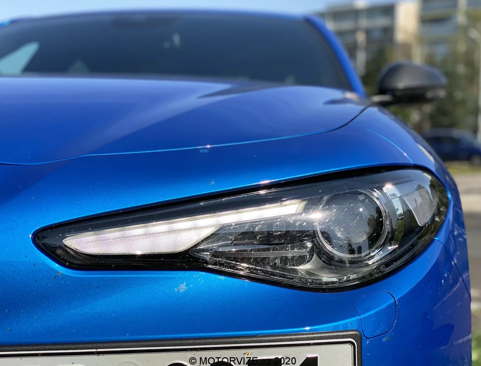 Primissimo piano del lato anteriore sinistro di una Alfa Romeo Giulia 2016 blu, catturato da un angolo basso che mostra i dettagli del faro e la curvatura del design dell'auto. Il faro moderno è dotato di illuminazione LED o HID con una forma angolare slanciata e un fascio principale di tipo proiettore per una luce focalizzata. Il cofano ha un design liscio e sagomato con una finitura metallica riflettente, e il paraurti anteriore presenta un design minimalista. Lo specchietto laterale, in tinta carrozzeria e aerodinamico, è parzialmente visibile nell'angolo in alto a destra, completando il profilo aerodinamico dell'auto.