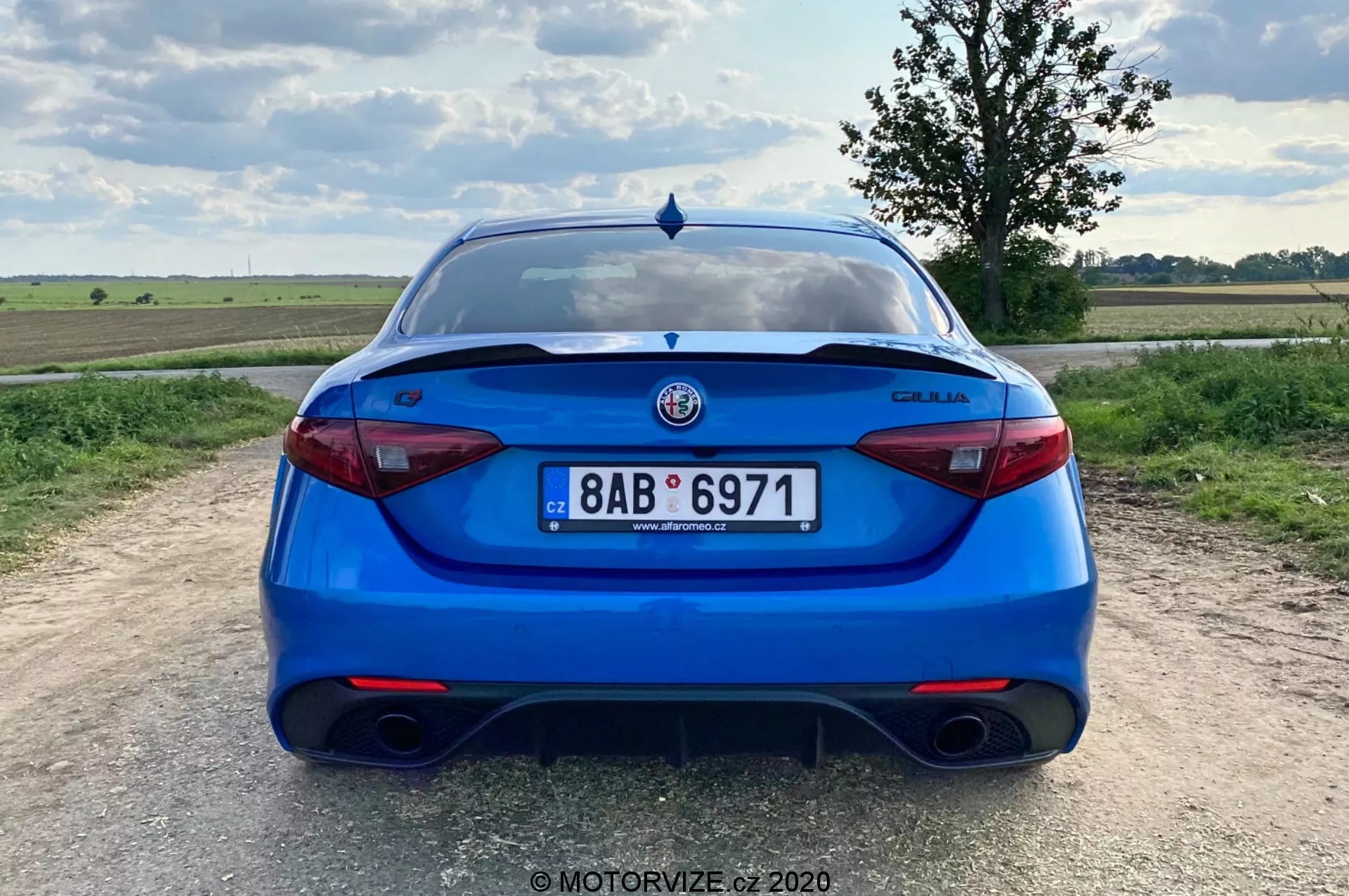 Vista posteriore di una berlina Alfa Romeo Giulia 2016 blu brillante, ripresa da una prospettiva a livello degli occhi. L'immagine mostra l'intera parte posteriore dell'auto, centrata nell'inquadratura, con la targa, le luci posteriori e il logo del produttore chiaramente visibili. Le caratteristiche includono un sistema di scarico doppio, luci posteriori moderne ed eleganti, un elemento del diffusore sul paraurti, uno spoiler sul portellone, sensori di parcheggio e un tergicristallo posteriore, suggerendo un equilibrio tra carattere sportivo, sicurezza e praticità. L'ambientazione è in una zona rurale con un paesaggio piatto e un cielo parzialmente nuvoloso durante il giorno.