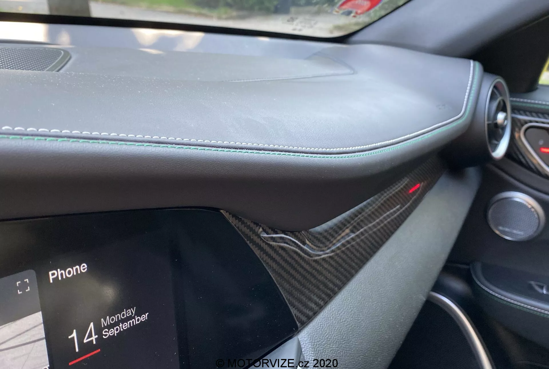 Primo piano degli interni della Alfa Romeo Giulia 2016 dalla prospettiva del passeggero, focalizzandosi sul cruscotto con materiale simile alla pelle e cuciture visibili, rifiniture simili al carbonio, schermo infotainment parzialmente visibile che mostra opzioni di connettività del telefono con il testo 'Phone' e 'Monday 14 September'. Luce rossa sopra il sistema infotainment che indica un sistema di sicurezza o di protezione attivo, enfatizzando lusso e design moderno.