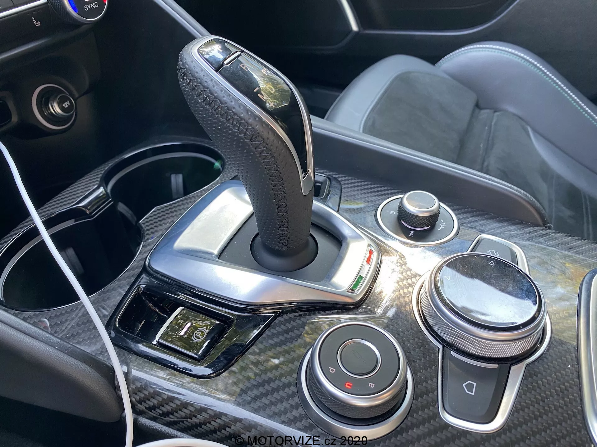 Vista ravvicinata della console centrale e del cambio dell'Alfa Romeo Giulia 2016 con leva del cambio automatico rivestita in pelle con finiture cromate, circondata da rifiniture in fibra di carbonio. La console comprende una manopola di controllo tipo iDrive, una manopola per il volume, pulsanti per la dinamica di guida, una porta USB con cavo collegato e un pannello con le luci di emergenza e altri controlli, sottolineando il design ergonomico e tecnologico degli interni dell'auto.