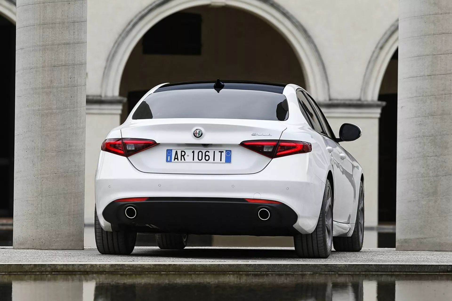 Alfa Romeo Giulia bianca del 2016 fotografata da un angolo posteriore di tre quarti che evidenzia il suo profilo posteriore sportivo, le luci posteriori distintive, i doppî scarichi e le caratteristiche di design elegante su sfondo di architettura europea