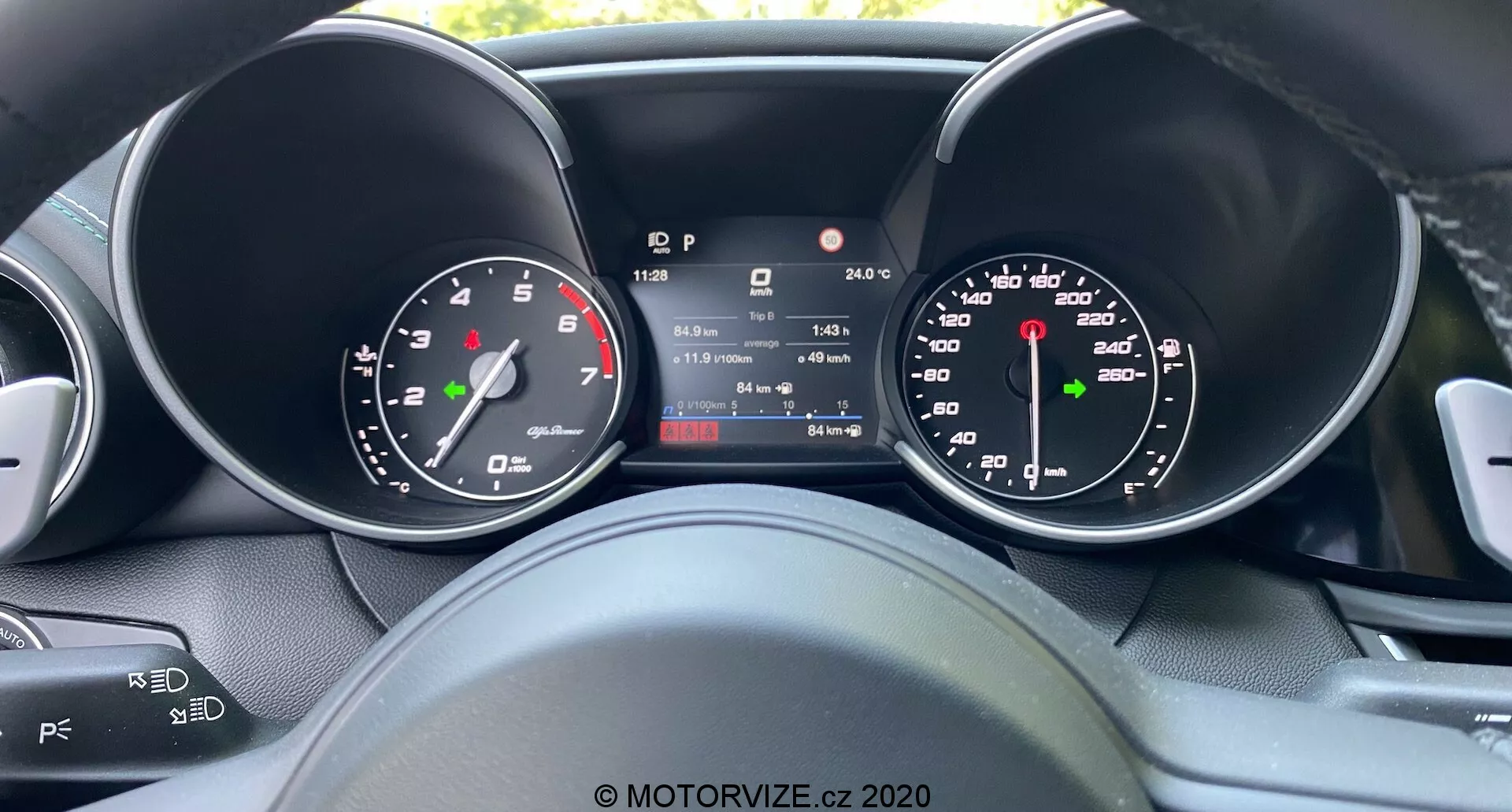 Visuale del conducente del quadro strumenti dell'Alfa Romeo Giulia 2016 con display digitale centrale che mostra velocità, marcia, temperatura e lettura del contachilometri, affiancato da tachimetro e contagiri analogici, che evidenzia una combinazione di design tradizionale e tecnologia moderna con viste parziali dei comandi al volante per l'audio e il cruise control.