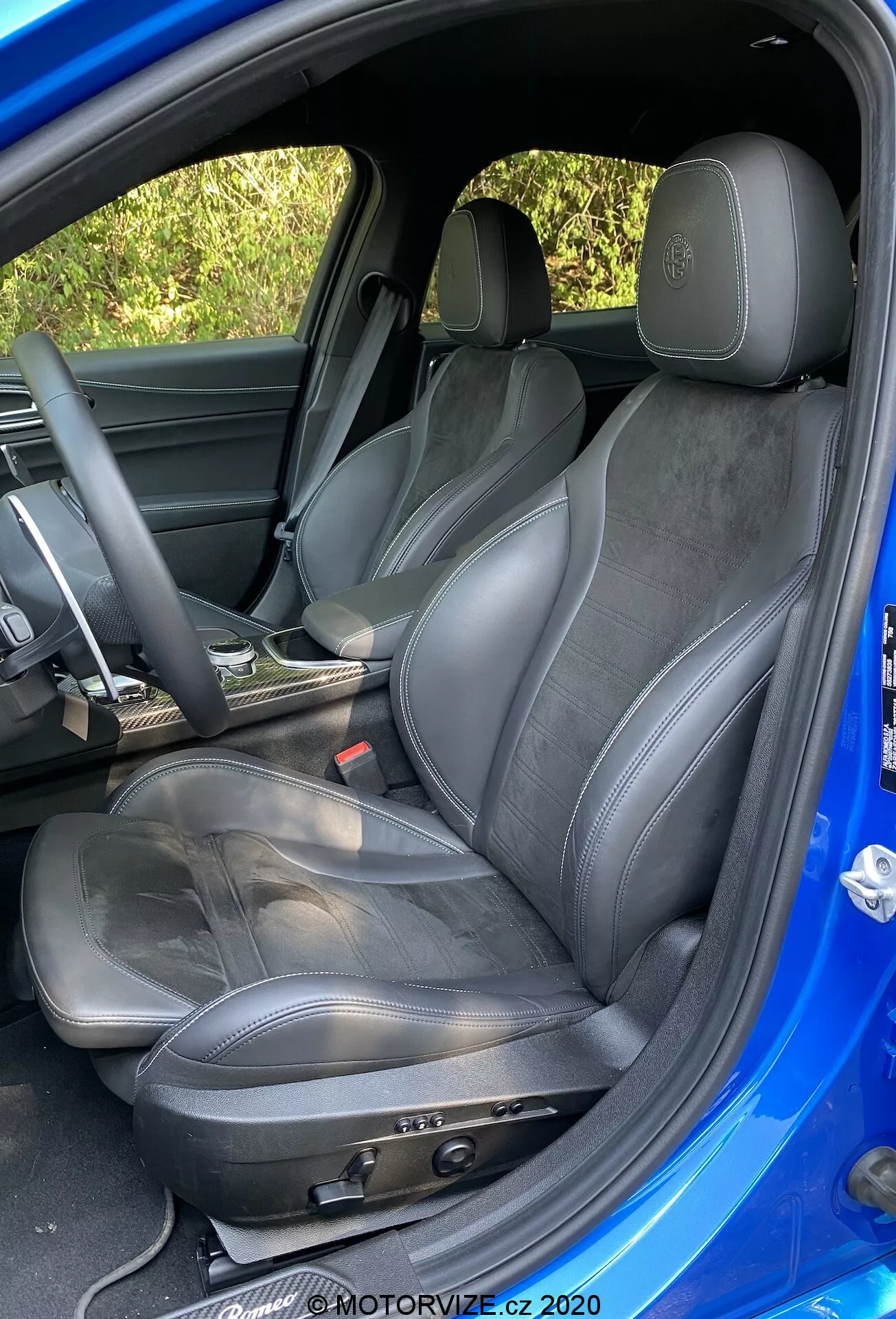 Vista dettagliata del sedile sportivo del conducente in una Alfa-Romeo Giulia 2016, che mostra regolazioni manuali del sedile, rivestimento in pelle e similpelle, logo in rilievo sul poggiatesta, leggero usura, volante parzialmente visibile rivestito in pelle e rivestimento della porta abbinato, catturato da un angolo esterno sopra il livello del sedile con interni scuri e esterni blu brillante a contrasto.