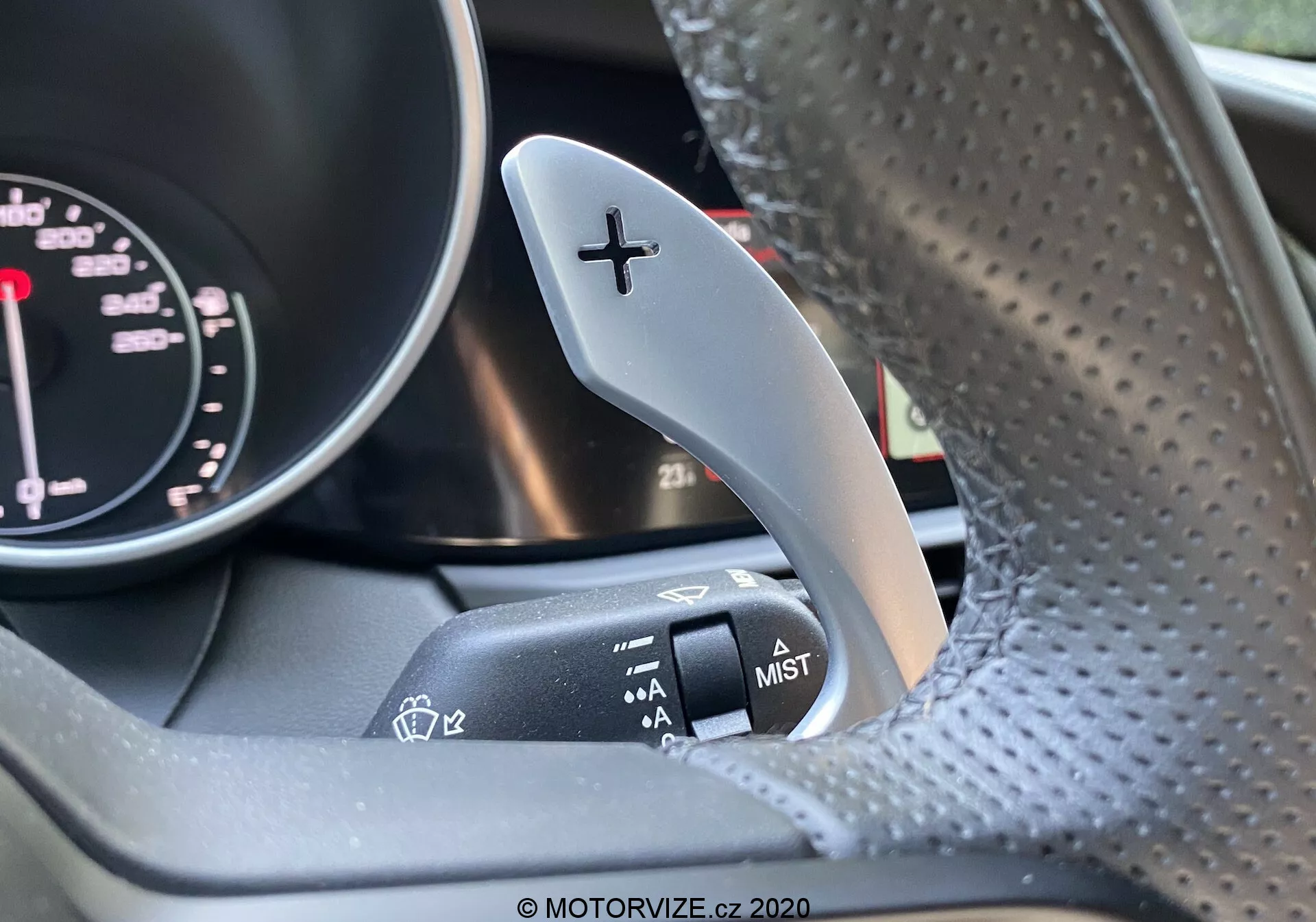 Primo piano dell'interno di un Alfa Romeo Giulia 2016, concentrandosi sulla leva del cambio a destra con il segno '+' per salire di marcia e sulla leva dei tergicristalli adiacente sulla colonna dello sterzo. Un lato del contagiri del quadro strumenti è leggermente visibile. Il volante, realizzato in pelle traforata, è leggermente sfocato. L'immagine evidenzia il contrasto tra la paletta del cambio in metallo e la finitura opaca della leva di controllo del tergicristallo.