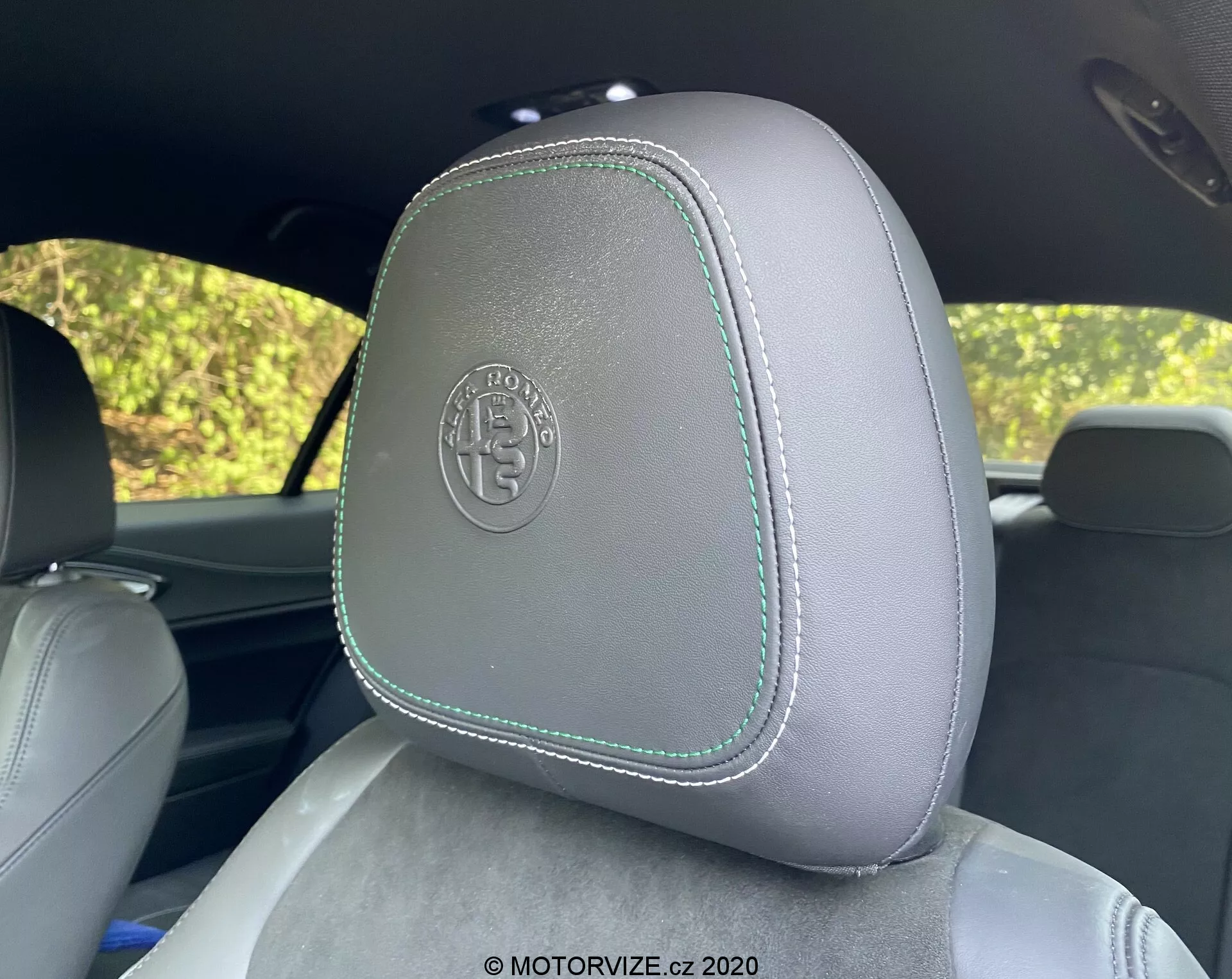 Primo piano di un poggiatesta auto nell'Alfa Romeo Giulia 2016, con rivestimento in pelle grigia chiara con cuciture a contrasto verde chiaro, visto dal sedile posteriore. Il poggiatesta presenta un logo circolare in rilievo che suggerisce una finitura premium o personalizzata. Lo sfondo mostra fogliame sfocato attraverso i finestrini dell'auto, suggerendo un ambiente esterno diurno.