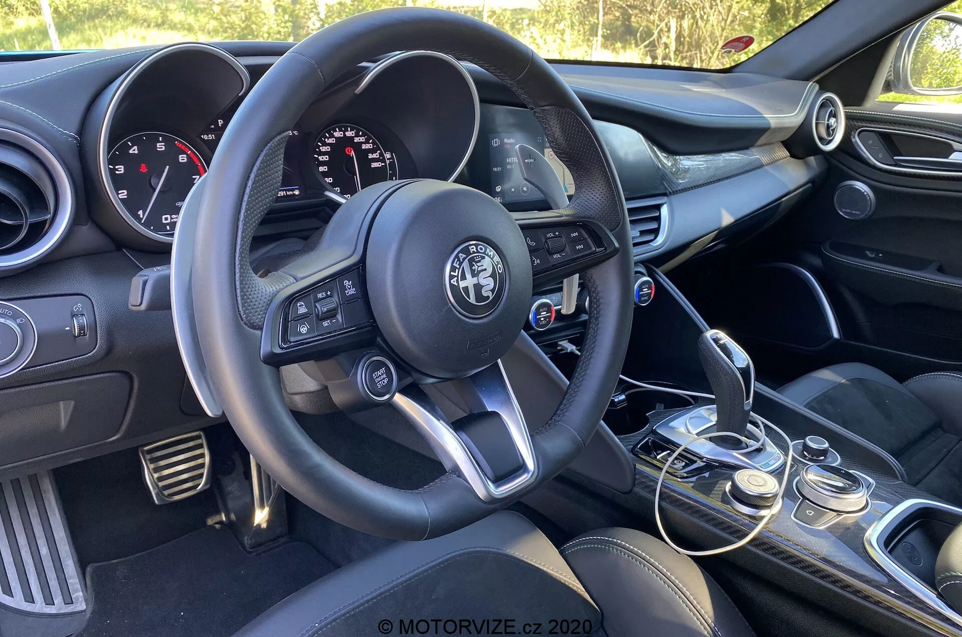 Vista interna dell'Alfa Romeo Giulia 2016 concentrata sul lato del conducente dalla prospettiva del passeggero, mostrando il volante a tre razze con pulsanti multifunzione, il logo del produttore al centro, il quadro strumenti con quadranti analogici e display digitale, la console centrale con comandi audio e climatizzazione, leva del cambio manuale con rivestimento in pelle, selettore delle modalità di guida, sedili anteriori con rivestimento scuro e pannello della portiera del conducente parzialmente visibile con comandi per finestrini e specchietti.