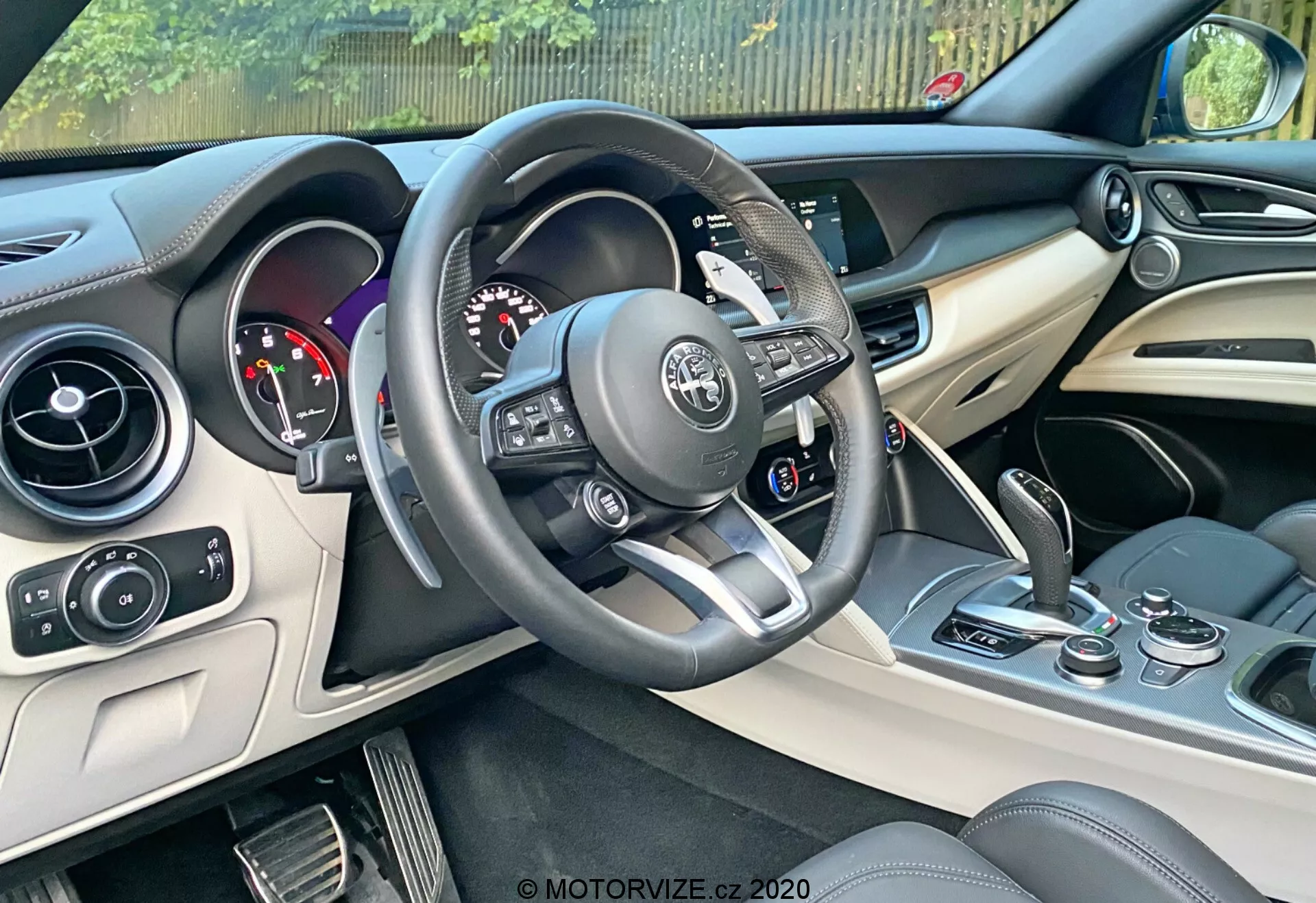 Immagine degli interni di un'Alfa Romeo Stelvio 2017 focalizzata sul lato del conducente. Presenta un volante multifunzionale rivestito in pelle con accenti in argento e controlli integrati, un quadro strumenti con quadranti analogici e display digitale, una console centrale con sistema di infotainment e controlli climatici, e uno sguardo ai sedili in pelle bicolore. Gli elementi di design moderno includono dettagli in argento o cromo sulla console e materiali lussuosi su cruscotto e porte. Include un pulsante di accensione/spegnimento vicino alla console centrale.