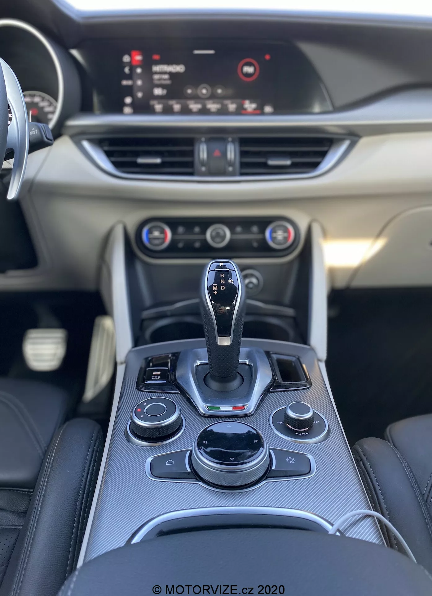 Vista dell'interno dell'Alfa Romeo Stelvio 2017, che mostra la console centrale con un cambio automatico e un array ergonomico di pulsanti e quadranti, inclusi infotainment, controlli del clima e forse modalità di guida. Il cruscotto presenta controlli aggiuntivi, prese d'aria e uno schermo infotainment in primo piano. Plancia lato passeggero liscia con finiture decorative. Fotografia di alta qualità che mette in risalto i materiali premium e il design elegante. © MOTORVIZE.cz