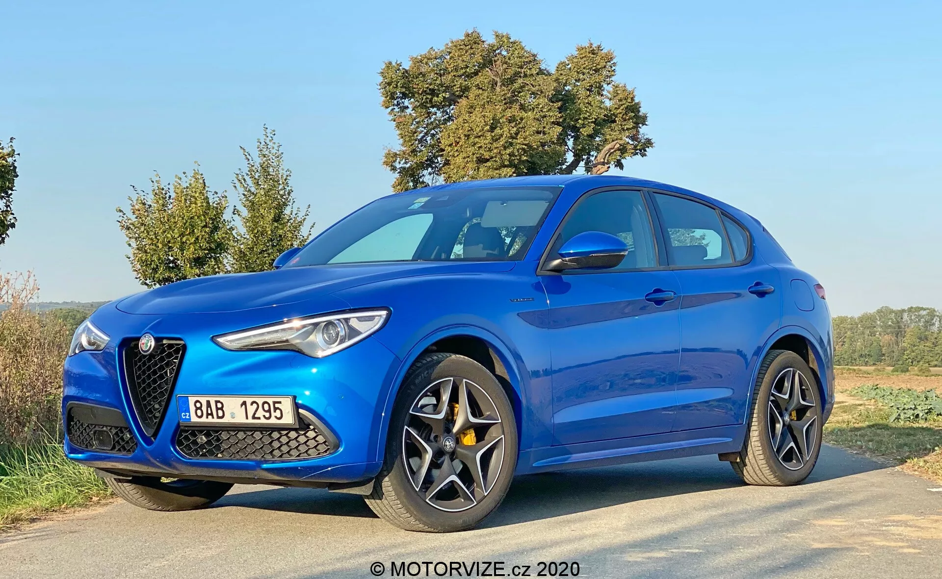 Alfa Romeo Stelvio 2017 blu, fotografato da un'angolazione frontale a tre quarti bassa, che mette in mostra la caratteristica griglia a forma di scudo, i fari allungati ed eleganti, le grandi prese d'aria, le linee sagomate del cofano e i cerchi in lega sportivi a più razze su una strada rurale.