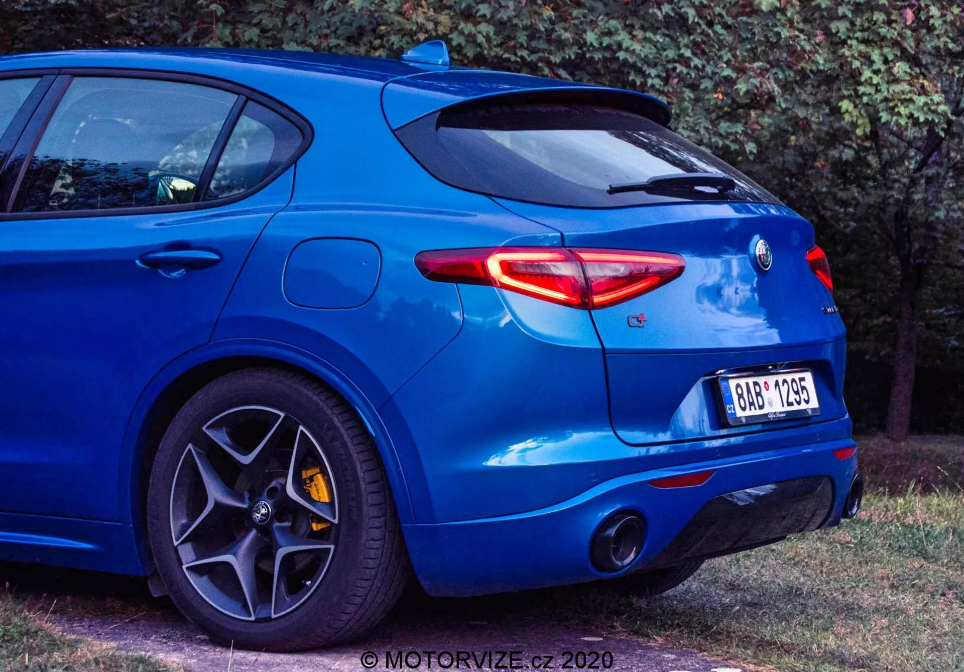Vista posteriore a tre quarti di una berlina sportiva blu Alfa Romeo Stelvio 2017, catturata da un'angolazione bassa per mettere in risalto le sue caratteristiche di design moderno, inclusi i sottili fari posteriori a LED, il sistema di scarico doppio, il badge posteriore centrato, lo spoiler posteriore montato sul tetto, i cerchi in lega a più razze con freni ad alte prestazioni, i pneumatici a basso profilo e la carrozzeria muscolosa con parafango posteriore sporgente. Il veicolo emana un'estetica sportiva e orientata alle prestazioni. Un watermark è visibile nell'angolo inferiore destro.
