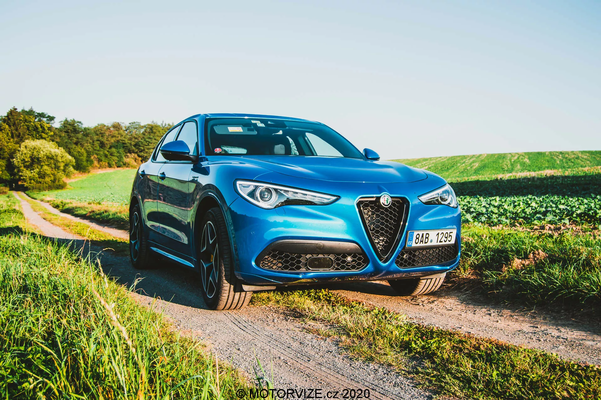 Alfa Romeo Stelvio 2017 blu parcheggiato su un sentiero di ghiaia accanto a un campo nel tardo pomeriggio, che mostra la griglia anteriore con contorno cromato, fari a LED, linee scolpite del cofano e cerchi in lega multirazze da un'angolazione frontale di tre quarti.
