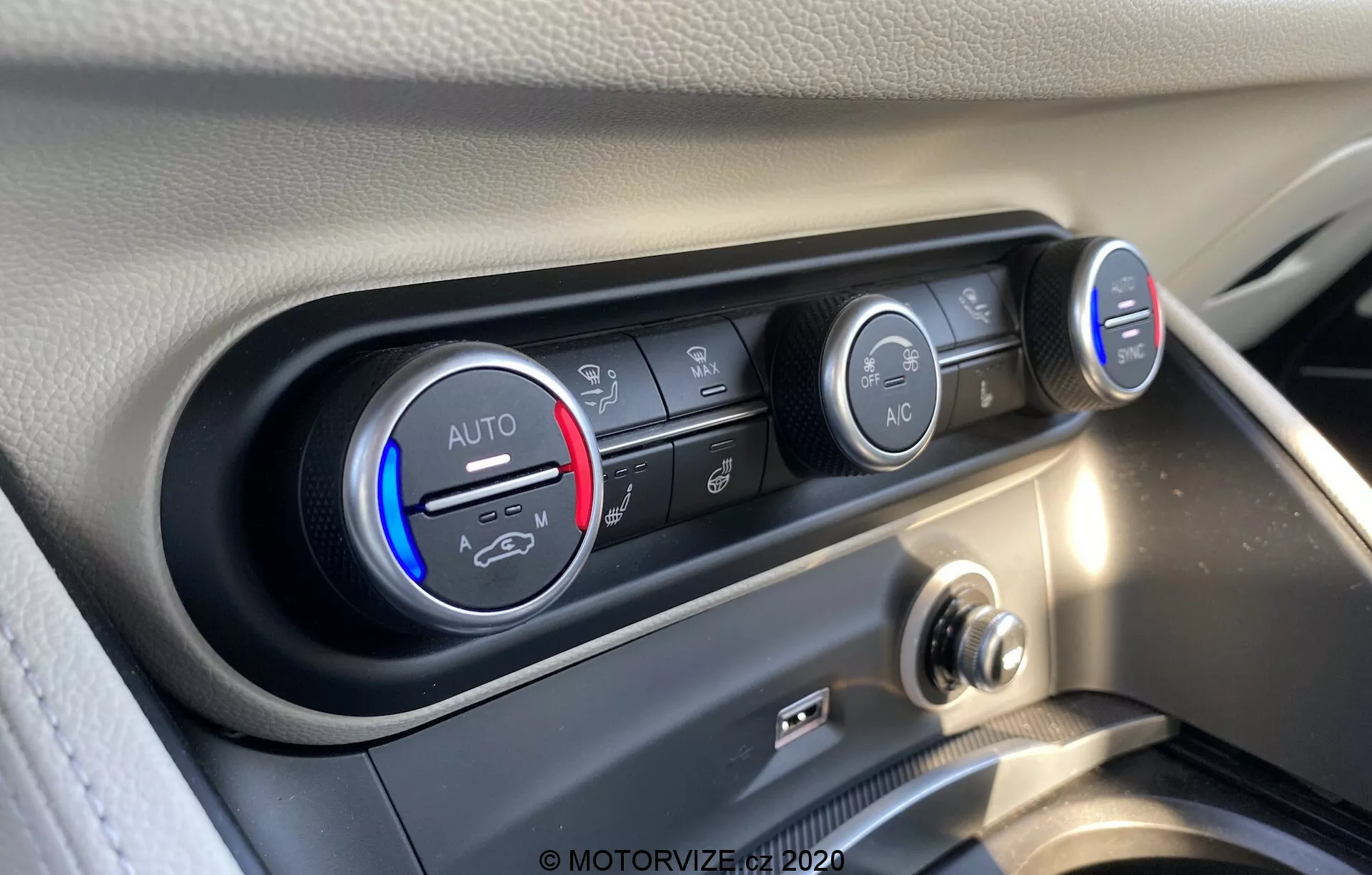 Vista ravvicinata del sistema di controllo del clima dell'interno dell'Alfa Romeo Stelvio 2017. L'immagine mostra le manopole e i pulsanti di controllo della temperatura e della ventola da un angolo leggermente elevato, mettendo a fuoco i dettagli. La manopola della temperatura, impostata su freddo, presenta una fascia blu e rossa con la scritta 'AUTO'. Accanto ci sono diversi pulsanti orizzontali per controllare la direzione del flusso d'aria, il ricircolo dell'aria e lo sbrinamento del parabrezza. La manopola destra indica la velocità della ventola con luci LED, impostata su un livello basso. Sotto spicca un pulsante 'A/C'. Il pannello di controllo è incastonato in una cornice nera lucida, in contrasto con il cruscotto chiaro, catturato ad alta risoluzione con chiari riflessi di luce naturale.