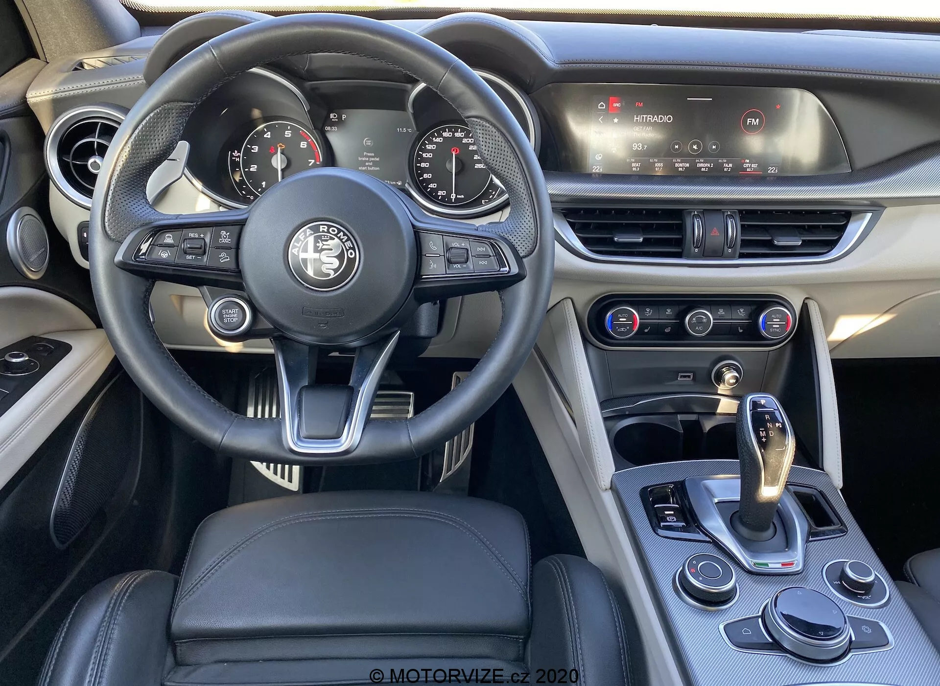 Scatto dell'interno di un Alfa Romeo Stelvio 2017, concentrato sull'area del conducente che mostra il volante multifunzione moderno rivestito in pelle con vari pulsanti di controllo, un quadro strumenti analogico-digitale parzialmente visibile e la console centrale con sistema di infotainment touchscreen e controllo del clima. Il design combina lusso e sportività con rivestimenti in pelle e cuciture a contrasto. La leva del cambio e il selettore delle modalità di guida evidenziano il cambio automatico e le modalità di guida selezionabili del veicolo.
