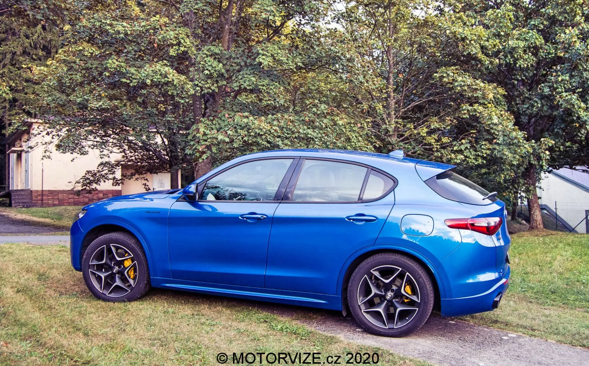 Vista di profilo laterale dell'Alfa Romeo Stelvio 2017 in blu brillante parcheggiato su un'area erbosa accanto a una superficie pavimentata, mostrando la vista anteriore con cerchi in lega neri multirazza, pinze dei freni gialle, specchi laterali in tinta carrozzeria, linea del tetto inclinata con antenna a pinna di squalo e design a quattro porte con finitura metallica.