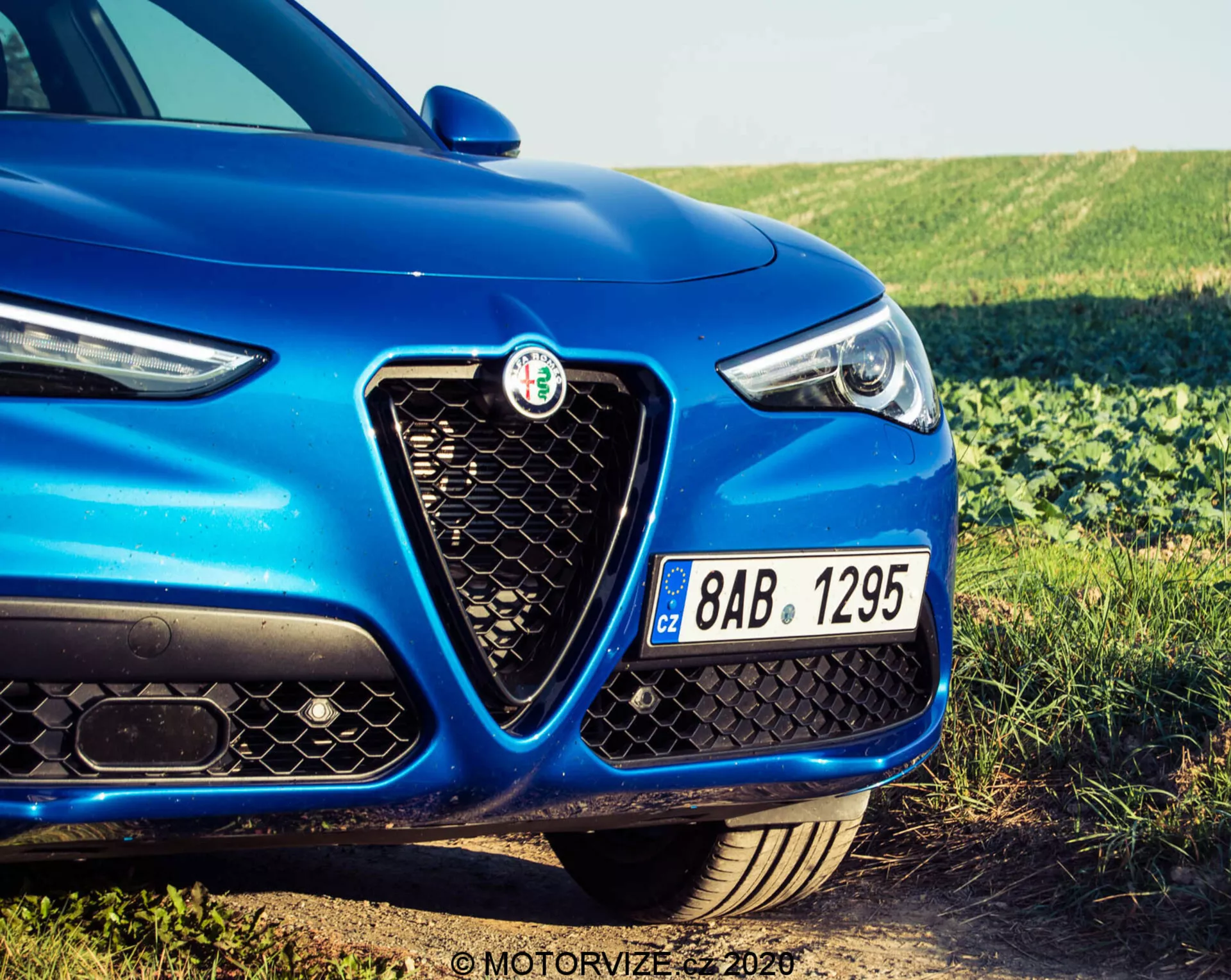 Primo piano del lato anteriore sinistro di un Alfa Romeo Stelvio 2017 blu, che mostra il distintivo badge sulla griglia esagonale, il design elegante dei fari con elementi LED e parte del cofano. L'immagine, scattata da un'angolazione bassa, mette in risalto la faccia anteriore sportiva e aggressiva del veicolo, compreso il paraurti anteriore nero con grandi prese d'aria e una targa ceca.