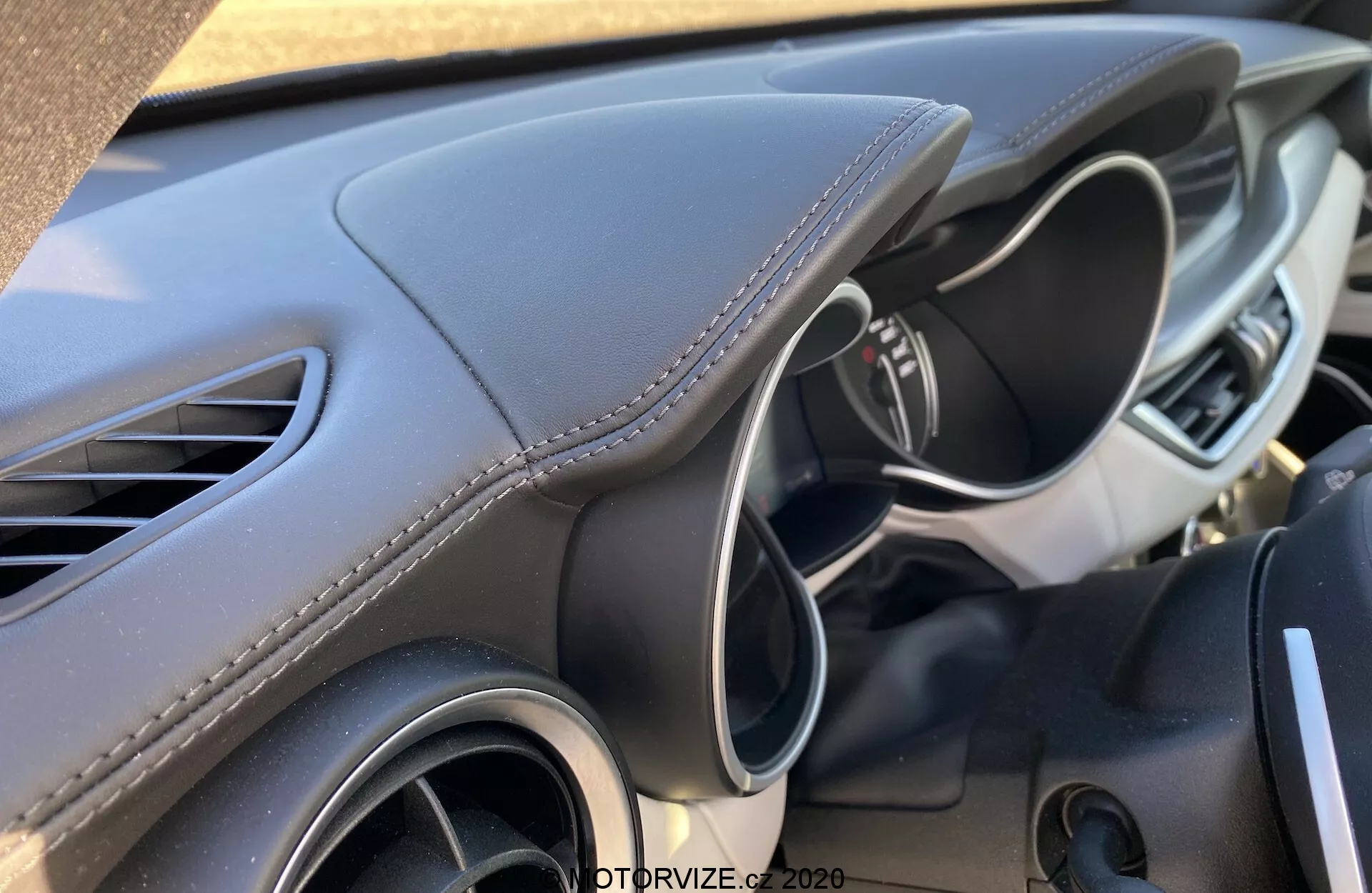 Vista ravvicinata degli interni dell'Alfa-Romeo Stelvio 2017 concentrata sul lato conducente del cruscotto, visibile dal sedile del passeggero o dalla consolle centrale. Il design moderno e scolpito del cruscotto presenta un materiale morbido al tatto con cuciture bianche, suggerendo un'estetica premium. Sono visibili anche la bocchetta d'aria sinistra e parte del volante multifunzione con comandi o pulsanti, suggerendo un ambiente di guida avanzato e facile da usare.