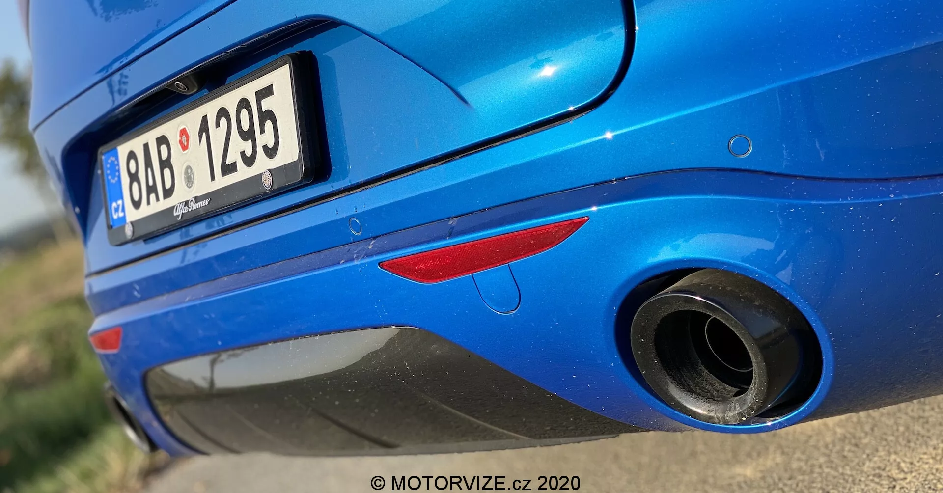Vista ravvicinata della sezione posteriore di una Alfa Romeo Stelvio 2017 blu, ripresa da un'angolazione bassa. L'inquadratura mette in evidenza la metà inferiore del retro del veicolo, focalizzandosi sul terminale di scarico e sul paraurti posteriore. Le caratteristiche includono una luce riflettente posteriore rossa, uno pneumatico posteriore parzialmente visibile, un paraurti posteriore arrotondato e lucido, una targa parzialmente ritagliata, un terminale di scarico cromato rotondo e una piega sopra il terminale di scarico sul paraurti. La vernice metallica con riflessi solari evidenzia le condizioni pulite e ben mantenute della vettura.