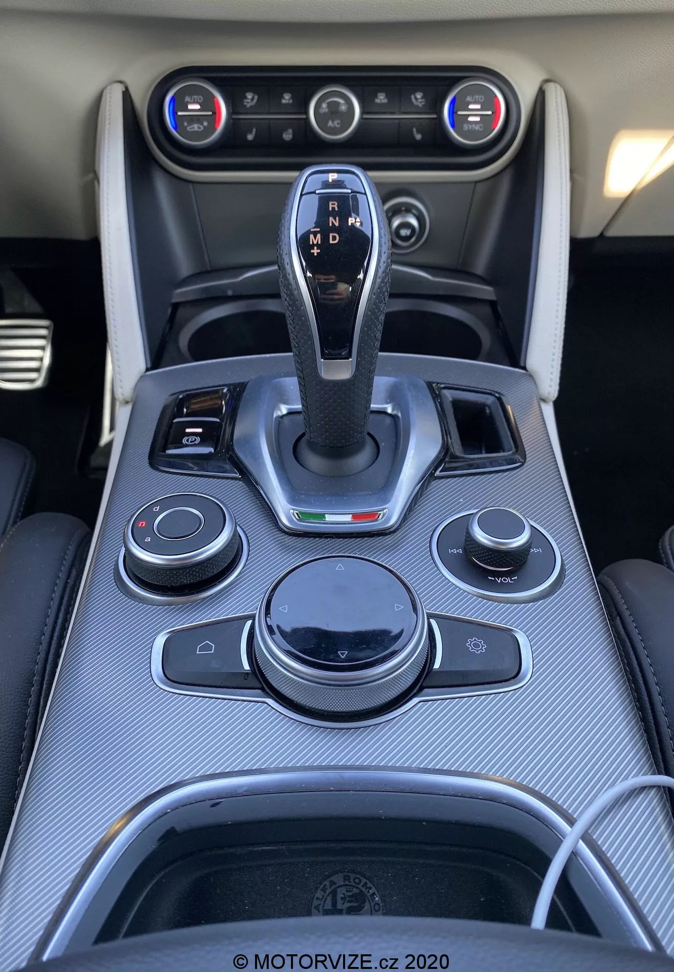 Vista ravvicinata dall'alto della console centrale nell'interno dell'Alfa Romeo Stelvio 2017. Leva del cambio centrale con finitura in pelle e accenti cromati, etichettata con impostazioni del cambio automatico. Manopole e pulsanti circostanti per sistema di infotainment, audio, clima e funzioni del veicolo. Rifinitura in fibra di carbonio, prese d'aria condizionata e sistema di climatizzazione a doppia zona visibili. Portabicchieri con contorno cromato e porta di connettività presente.