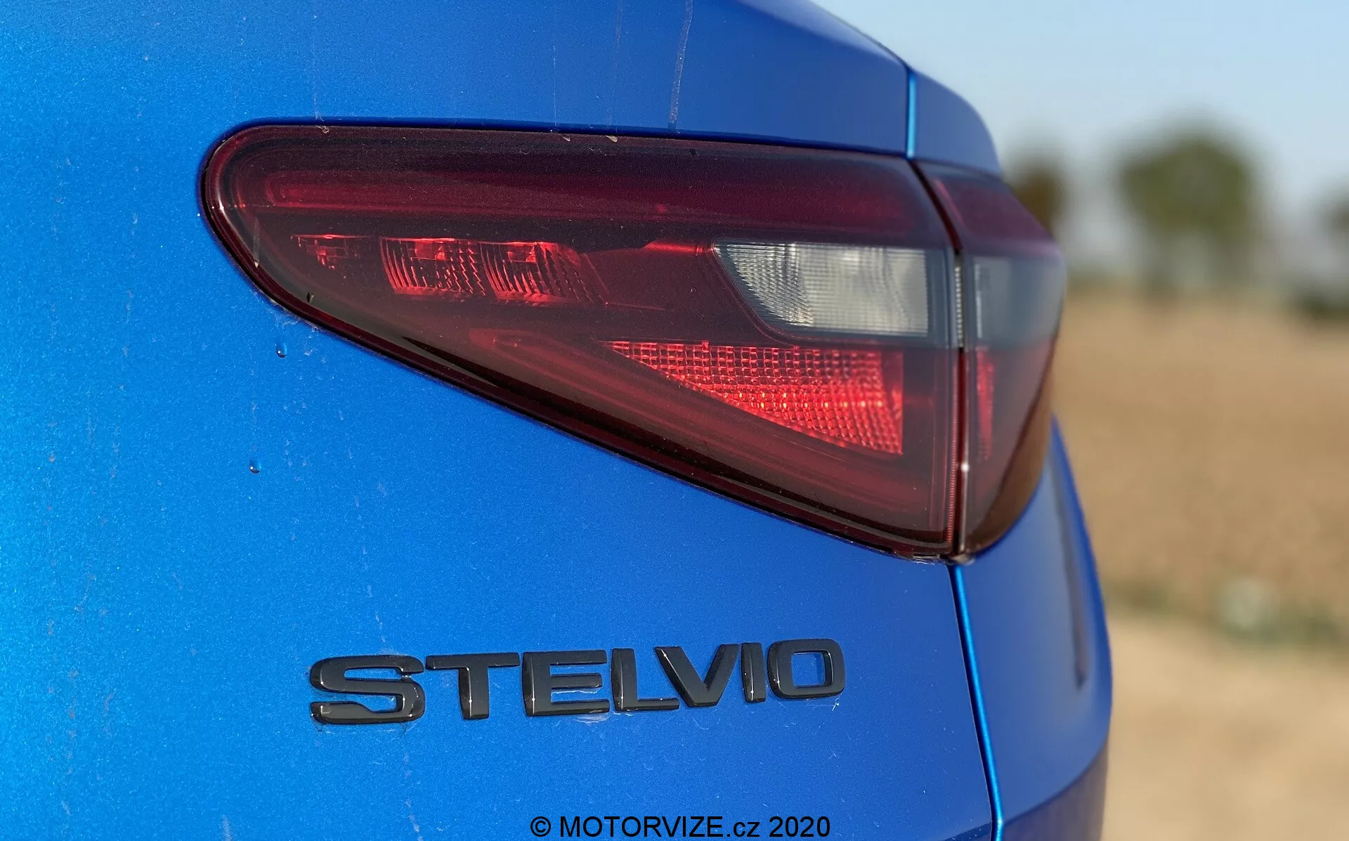 Dettaglio della sezione posteriore dell'Alfa Romeo Stelvio 2017 blu, catturato da un'angolazione bassa e leggermente fuori centro, focalizzandosi sul lato posteriore sinistro. Evidenzia il moderno fanale posteriore a LED e il badge cromato 'STELVIO', mostrando la finitura della vernice metallica distintiva. La bassa profondità di campo mantiene il fanale posteriore e il badge a fuoco contro uno sfondo sfocato. La luce naturale del giorno esalta la profondità del colore e i riflessi.