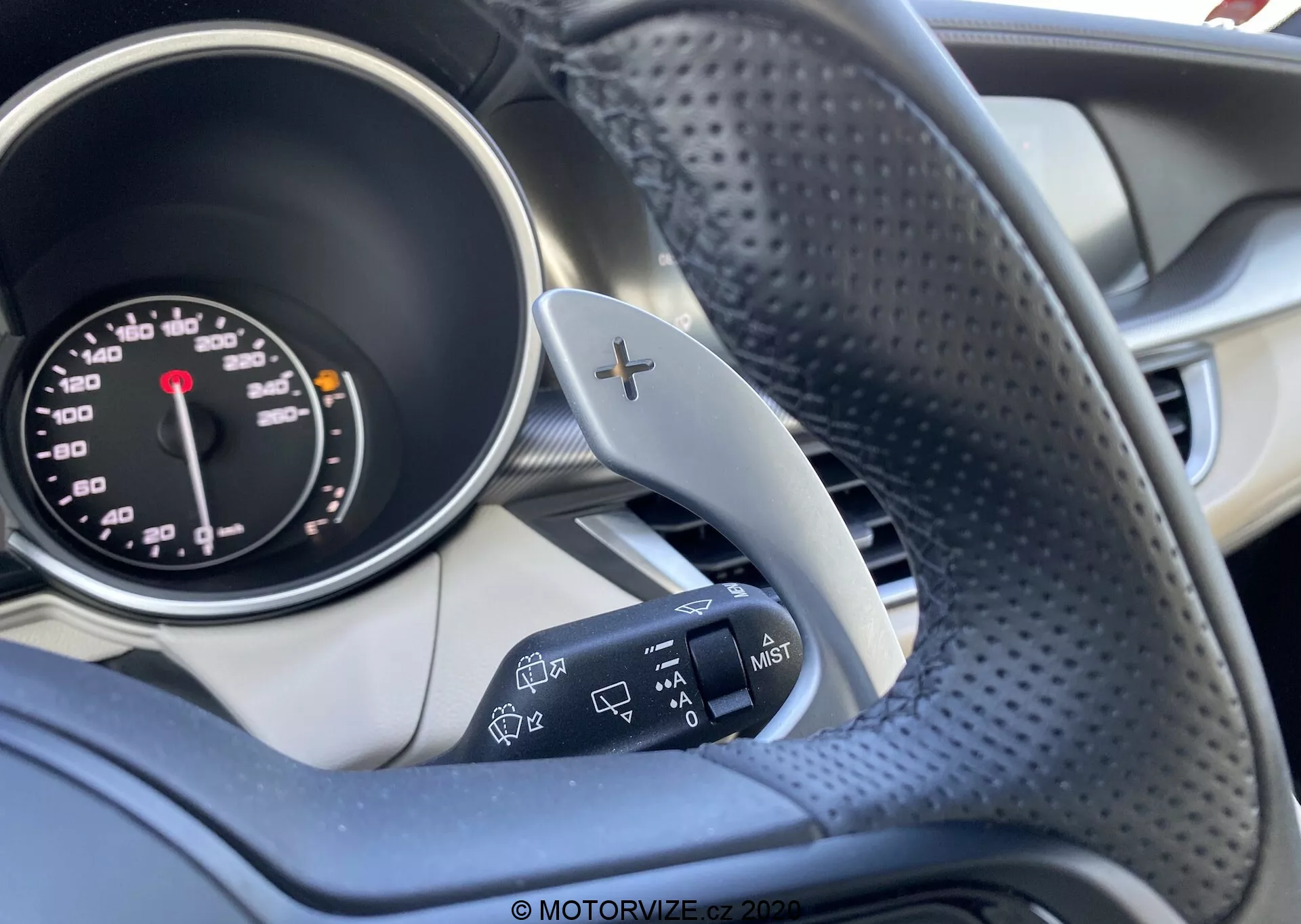 L'immagine mette in evidenza l'interno di un'Alfa Romeo Stelvio 2017, concentrandosi sul lato del conducente con un'enfasi sul volante e parte del quadro strumenti. La foto è scattata dal lato destro del volante, mostrando la parte sinistra del volante con trama in pelle traforata e un accento metallico con un simbolo '+' che indica i paddle shifters. Si vede anche il tachimetro analogico con le unità in mph e km/h e la leva dell'indicatore di direzione sulla sinistra con comandi aggiuntivi per i fari. Lo sfondo presenta elementi interni sfocati con una combinazione di toni scuri e chiari. L'attenzione ai dettagli sui comandi mette in risalto la funzionalità di sterzo e di illuminazione del veicolo.
